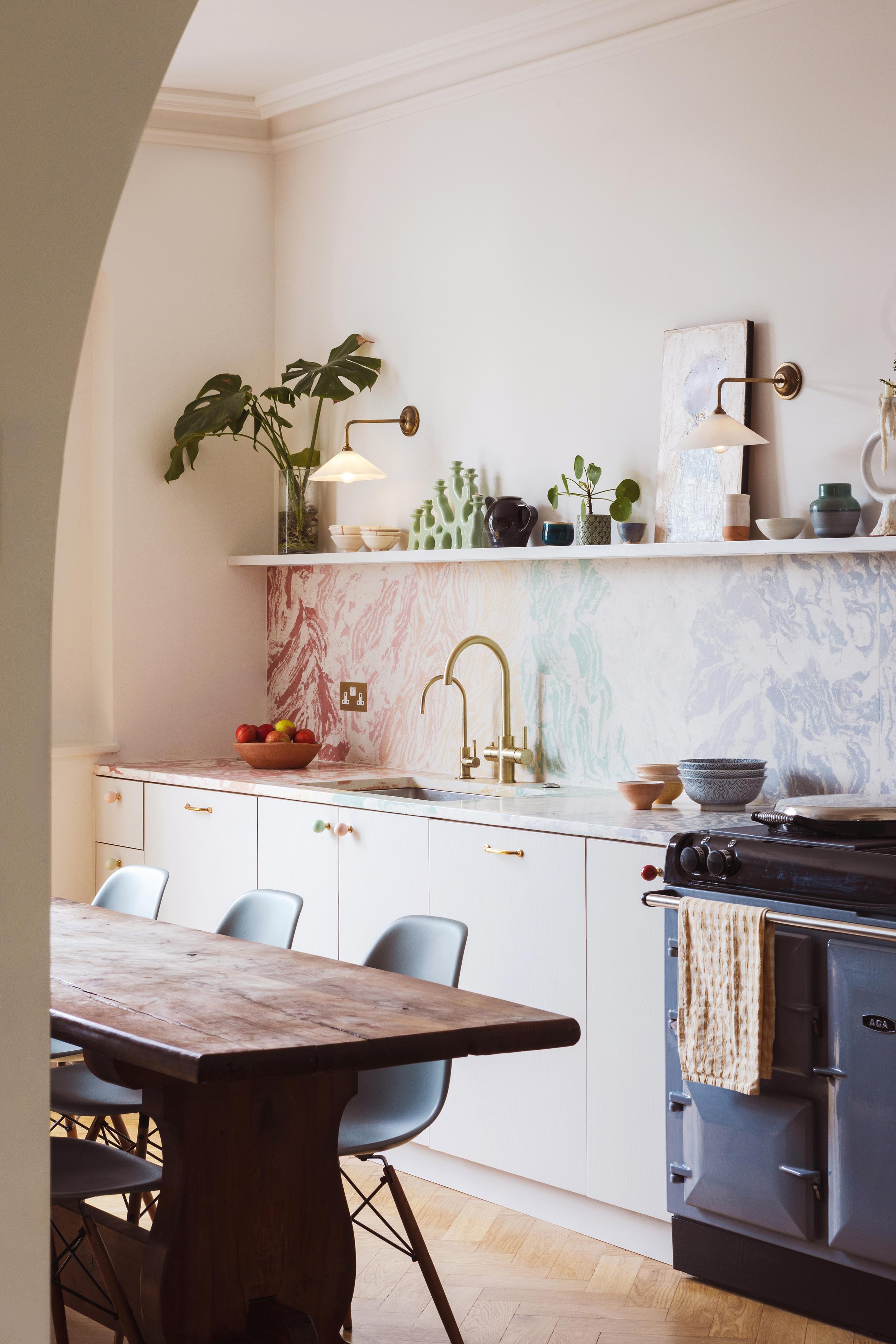A cozy kitchen with a wooden dining table and light blue chairs. The countertop features a marble backsplash, brass faucet, plants, and dishes. An archway frames the view, and natural light illuminates the room.