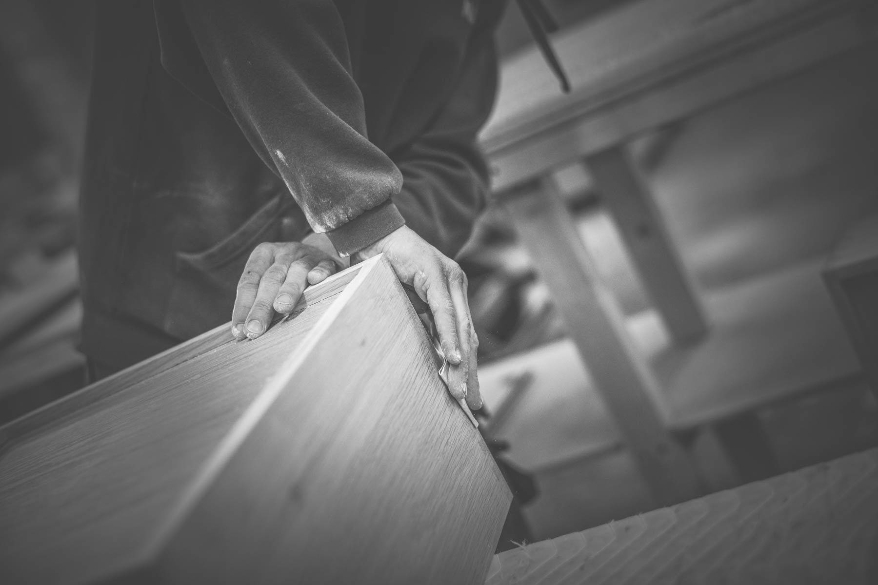 A person wearing a long-sleeve shirt uses both hands to work on a wooden piece in a workshop. The image is in black and white, emphasizing the focus on the craftsmanship and texture of the wood.