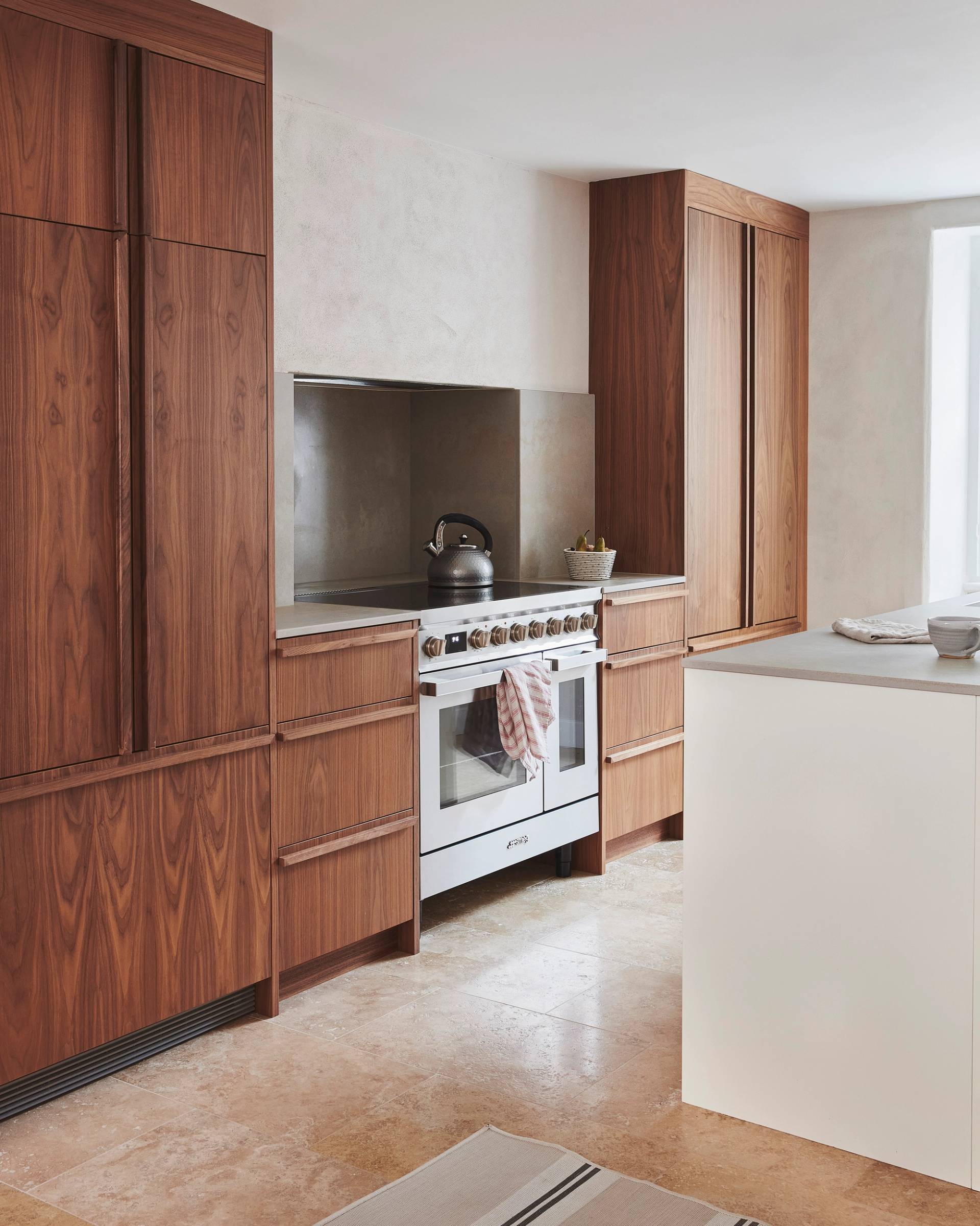 Modern kitchen with wooden cabinets, a white stove with a kettle on top, and a towel draped over the oven handle. A white countertop island is in the foreground, and a window allows natural light to fill the space.