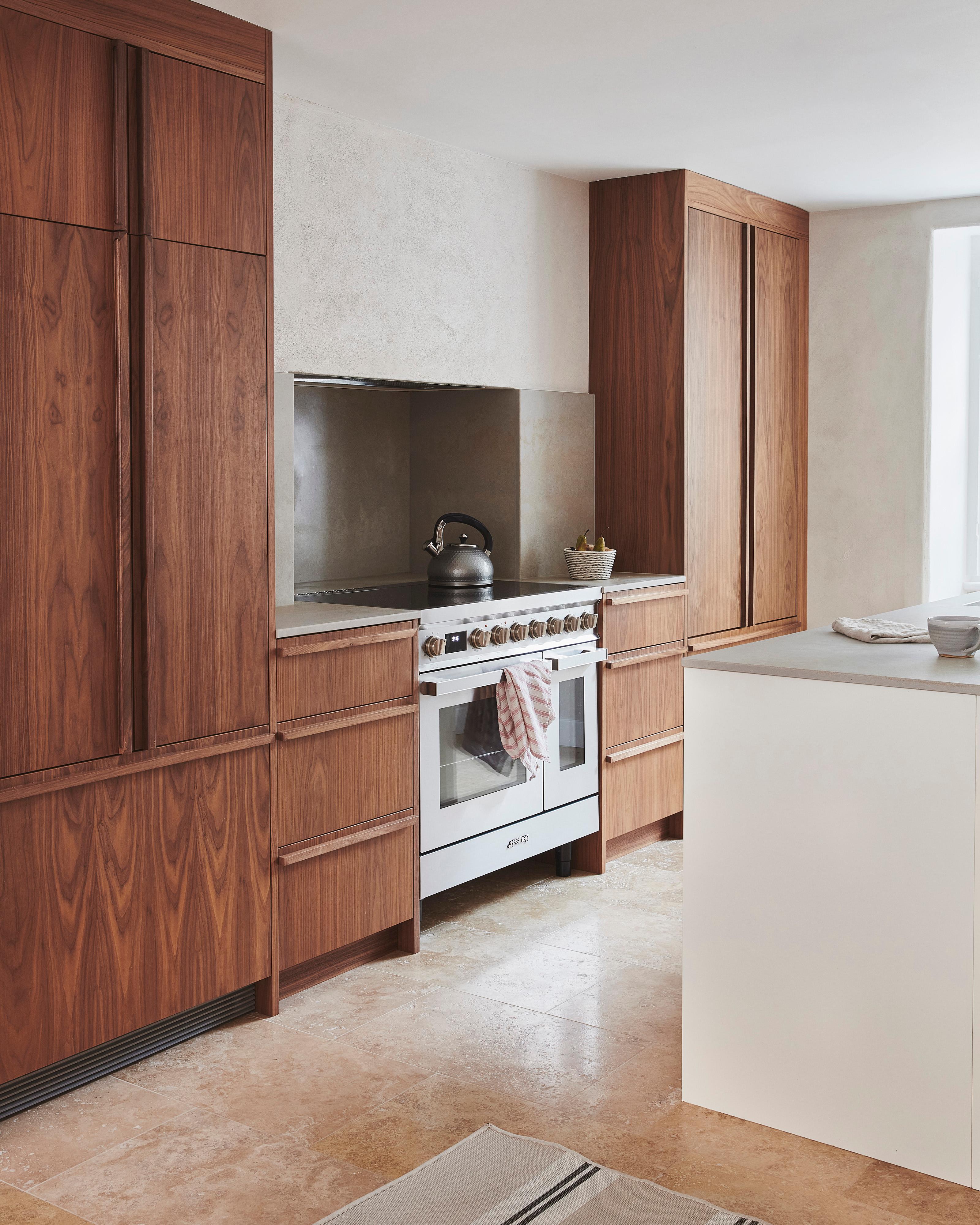 Modern kitchen with wooden cabinets, a white stove with a kettle on top, and a towel draped over the oven handle. A white countertop island is in the foreground, and a window allows natural light to fill the space.