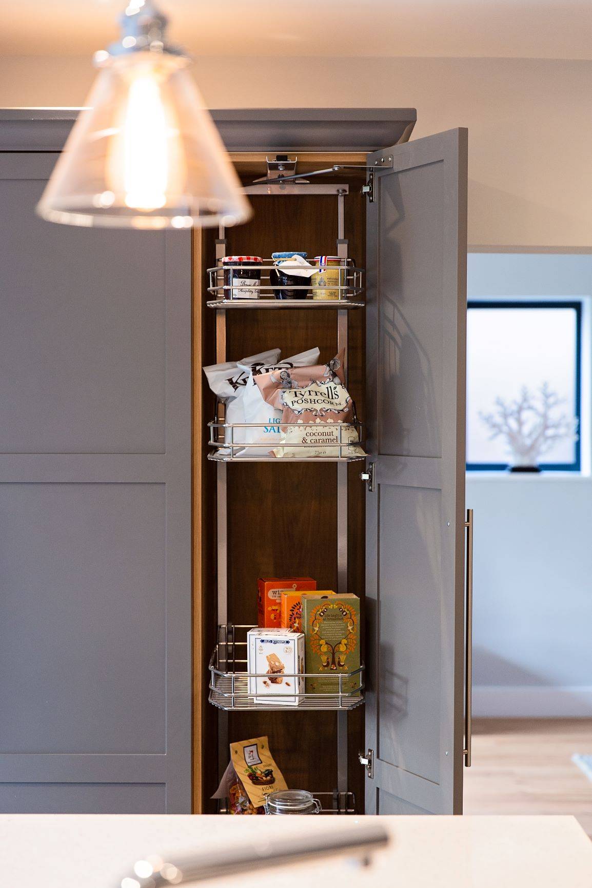 Grey kitchen cabinet with a pull-out pantry shelves opened, showcasing various snack items including popcorn, cracker boxes, and tea packets. A pendant light hangs above, and a window in the background provides natural light.