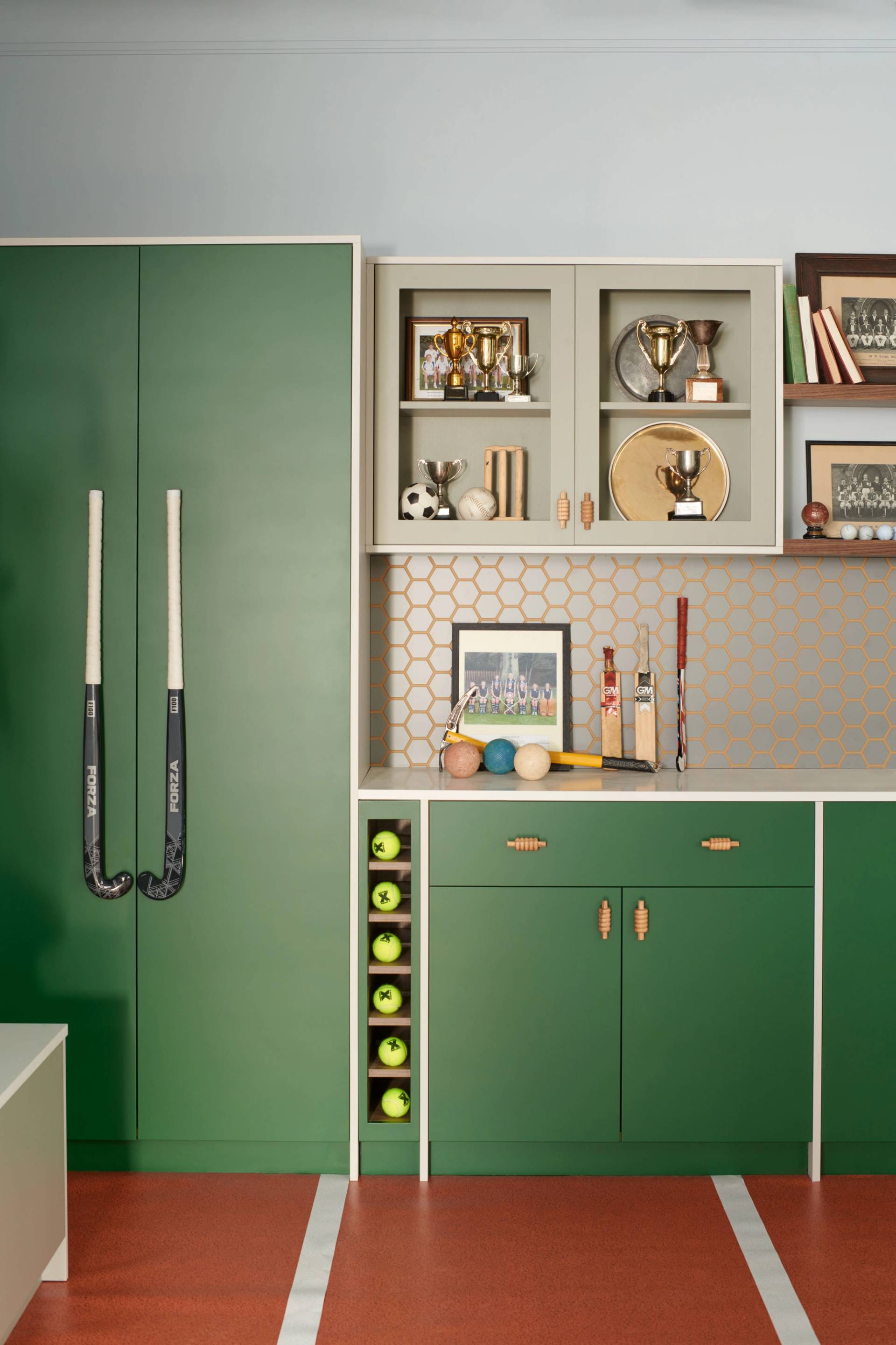 A detailed shot of a sports-themed interior with a vibrant green and orange color scheme. Forest green cabinets feature long black field hockey sticks as door handles, while a vertical rack holds a row of bright yellow tennis balls. Above the white countertop, a grey wall is decorated with a gold honeycomb pattern. Display cabinets and wooden shelves showcase vintage sports memorabilia, including gold trophies, cricket bats, and framed team photographs. The floor is a burnt orange color with white lines mimicking a sports court.