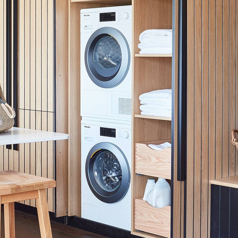 A modern laundry nook with a stacked washer and dryer, wooden shelves holding neatly folded white towels, and two drawers containing bottles, set against vertical wood paneling.