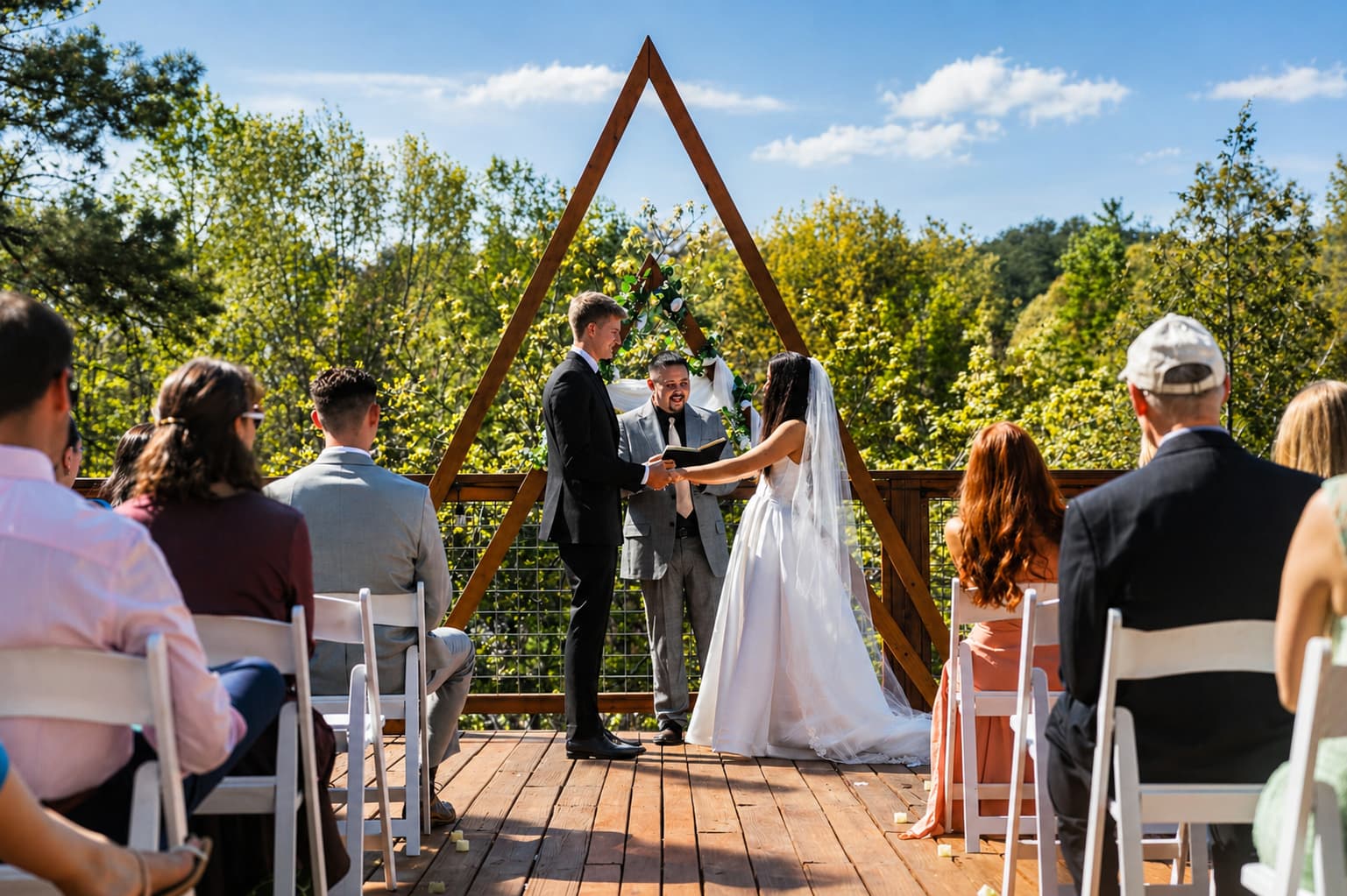 Couple standing under a wooden arch with guests and trees surrounding them.