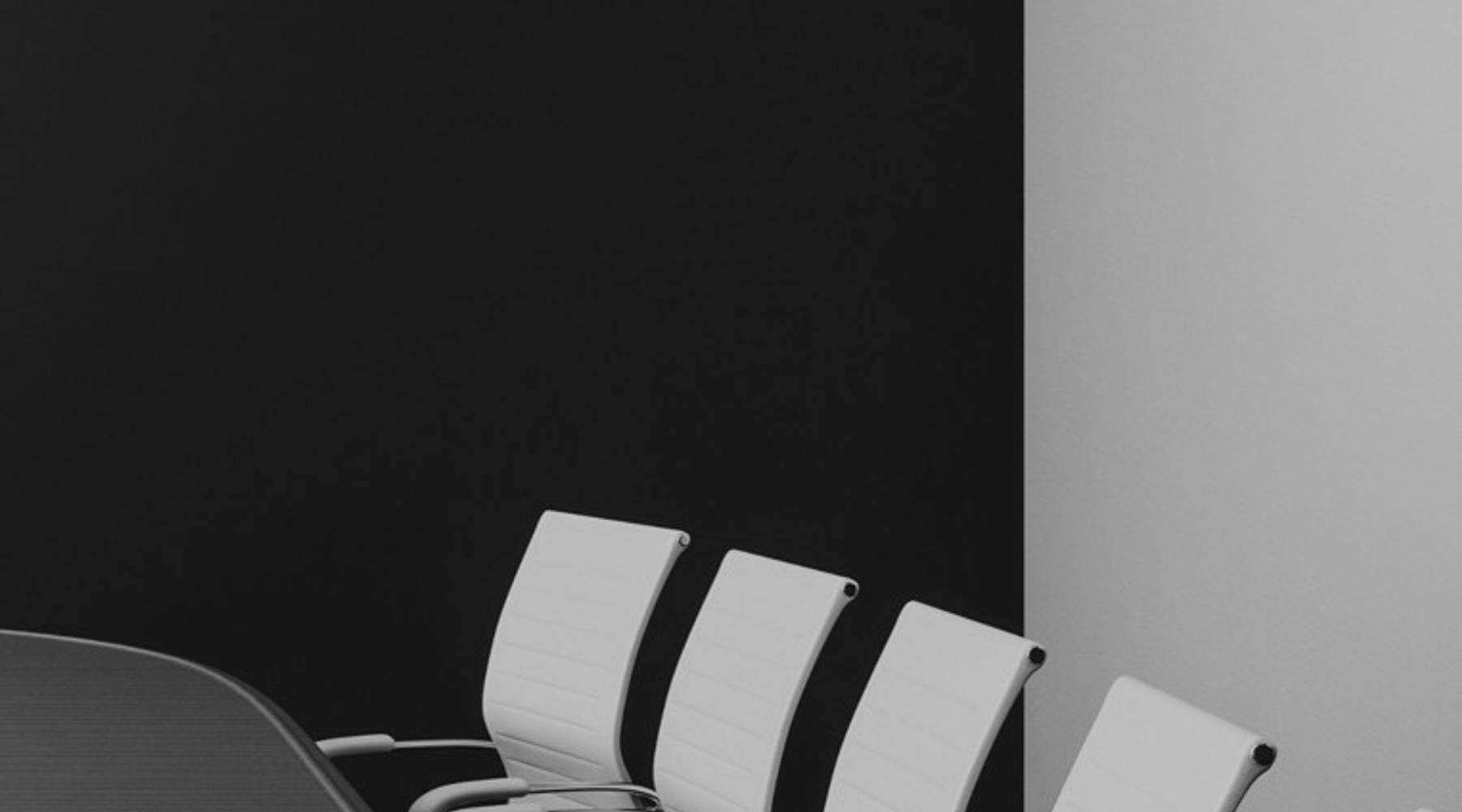 Photograph of a modern conference room with a long meeting table and a row of white office chairs aligned along the side, set against a dark wall. The layout conveys a professional business environment.