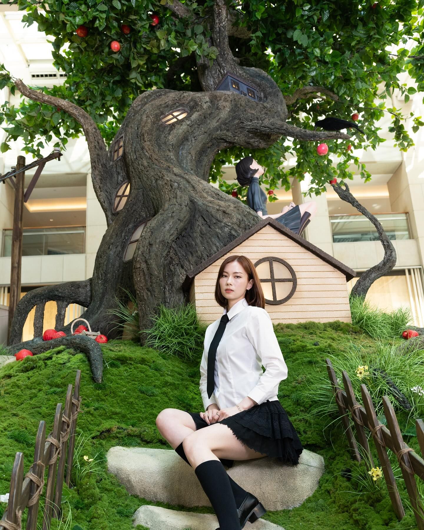Woman in a white shirt and black skirt sits before a whimsical apple tree with tiny windows and a cottage.