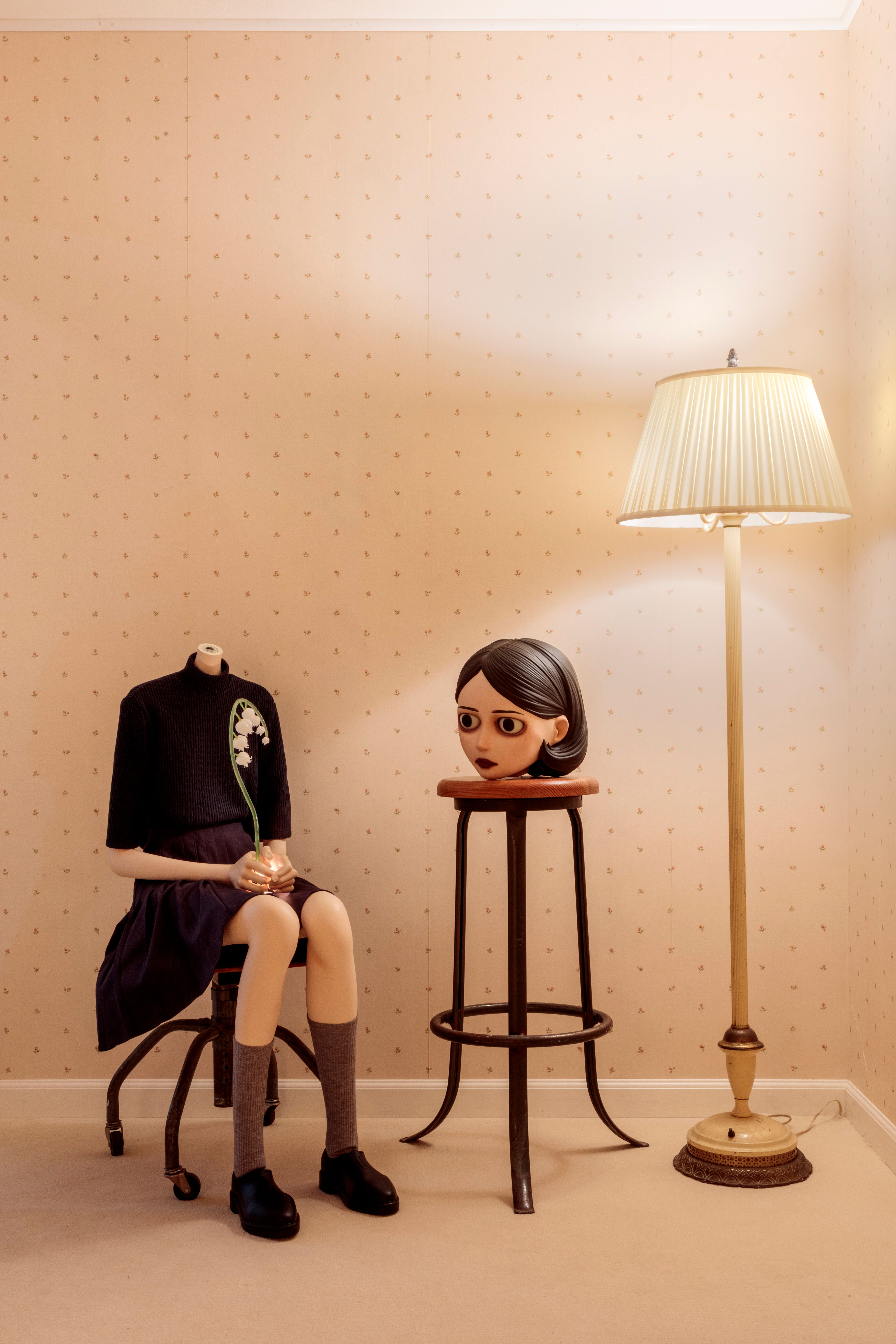 Surreal sculpture of a seated headless girl holding a flower, with her head on a stool beside a floor lamp.