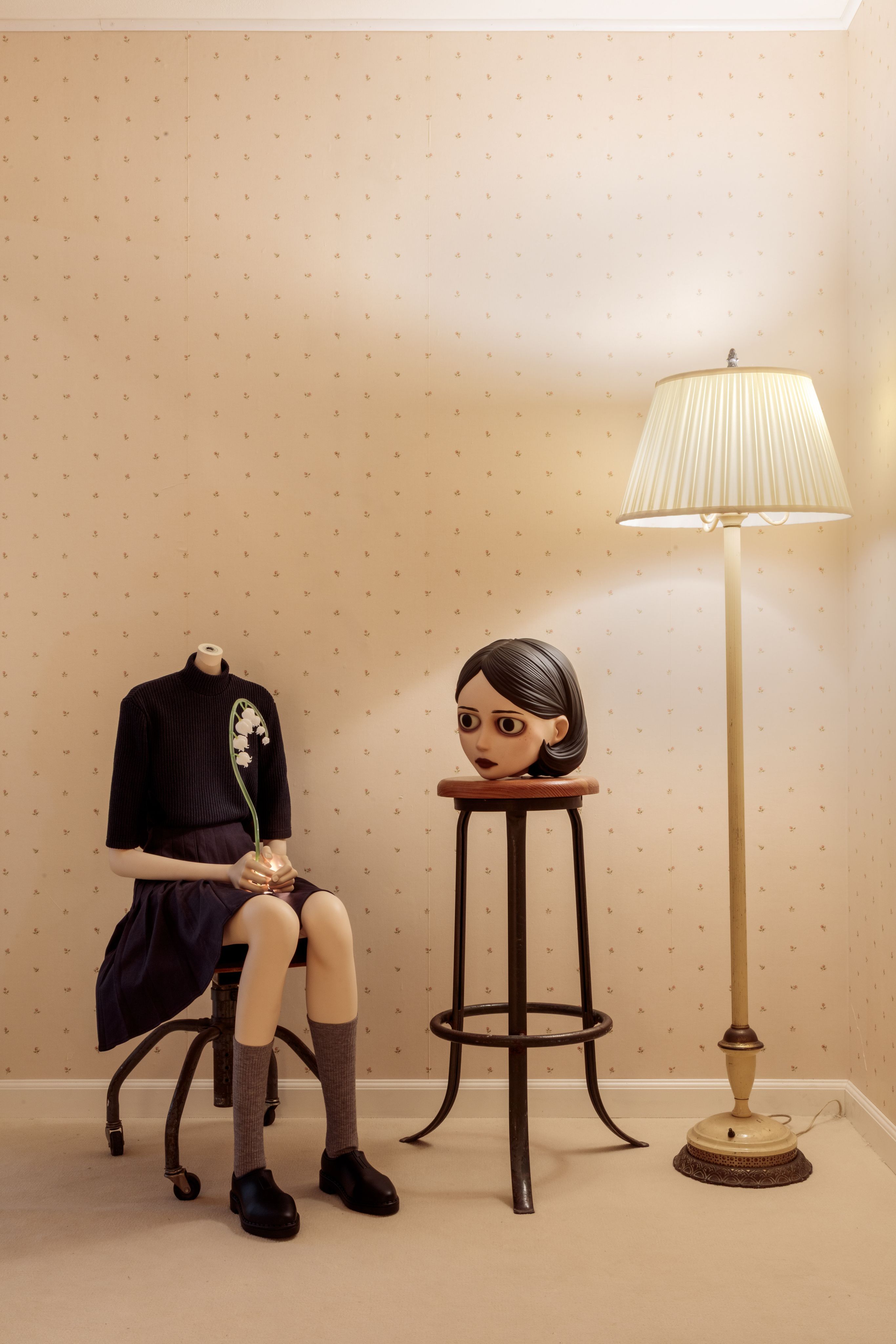 Surreal sculpture of a headless seated girl holding a flower, her head on a stool beside a floor lamp