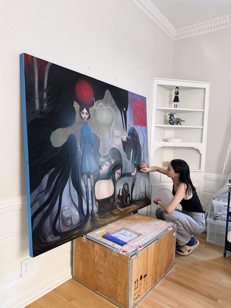 Artist crouches beside a large surreal figurative painting propped above a wooden shipping crate.