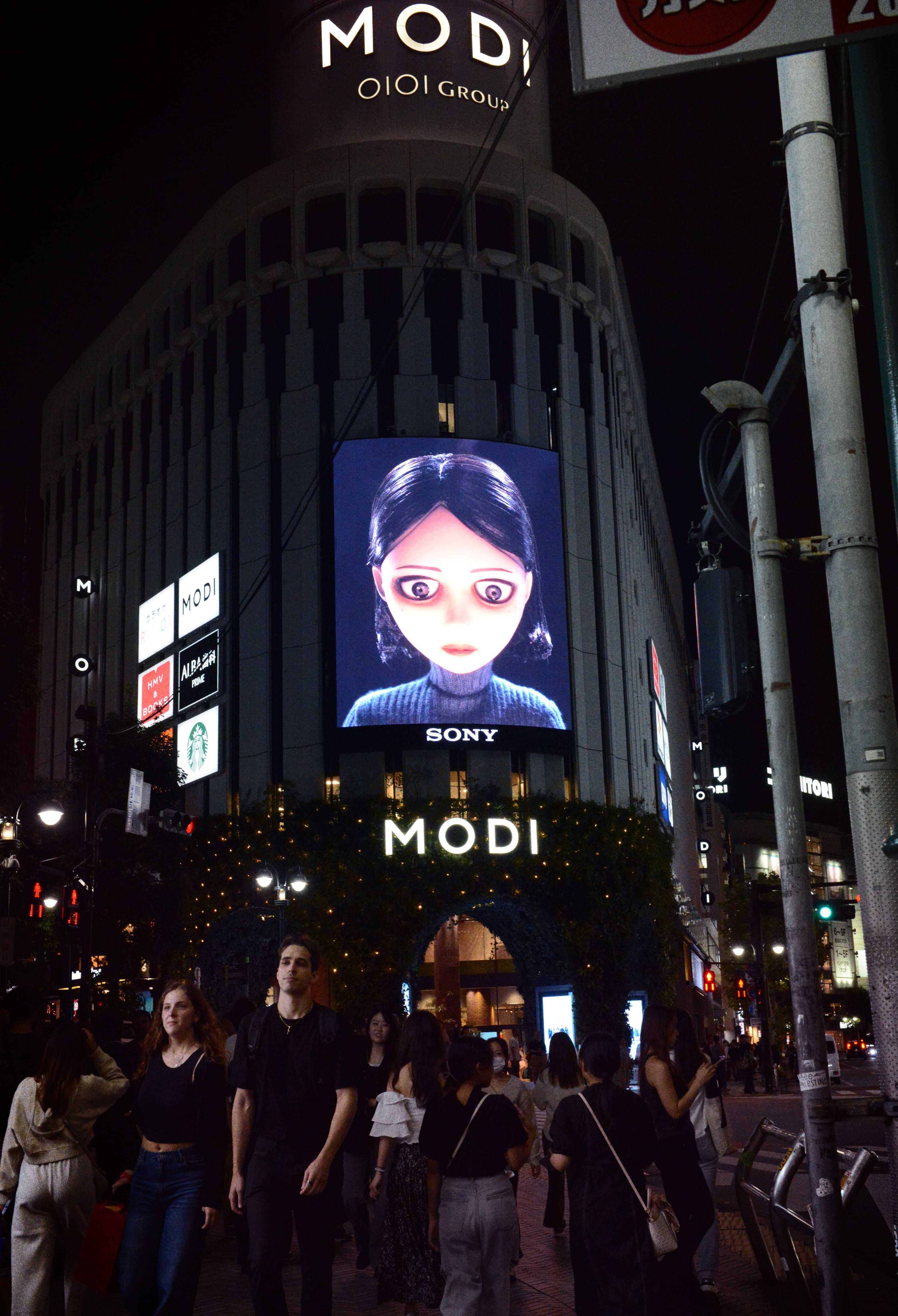 Crowds pass Tokyo's MODI building at night beneath a Sony screen showing an animated girl's face.