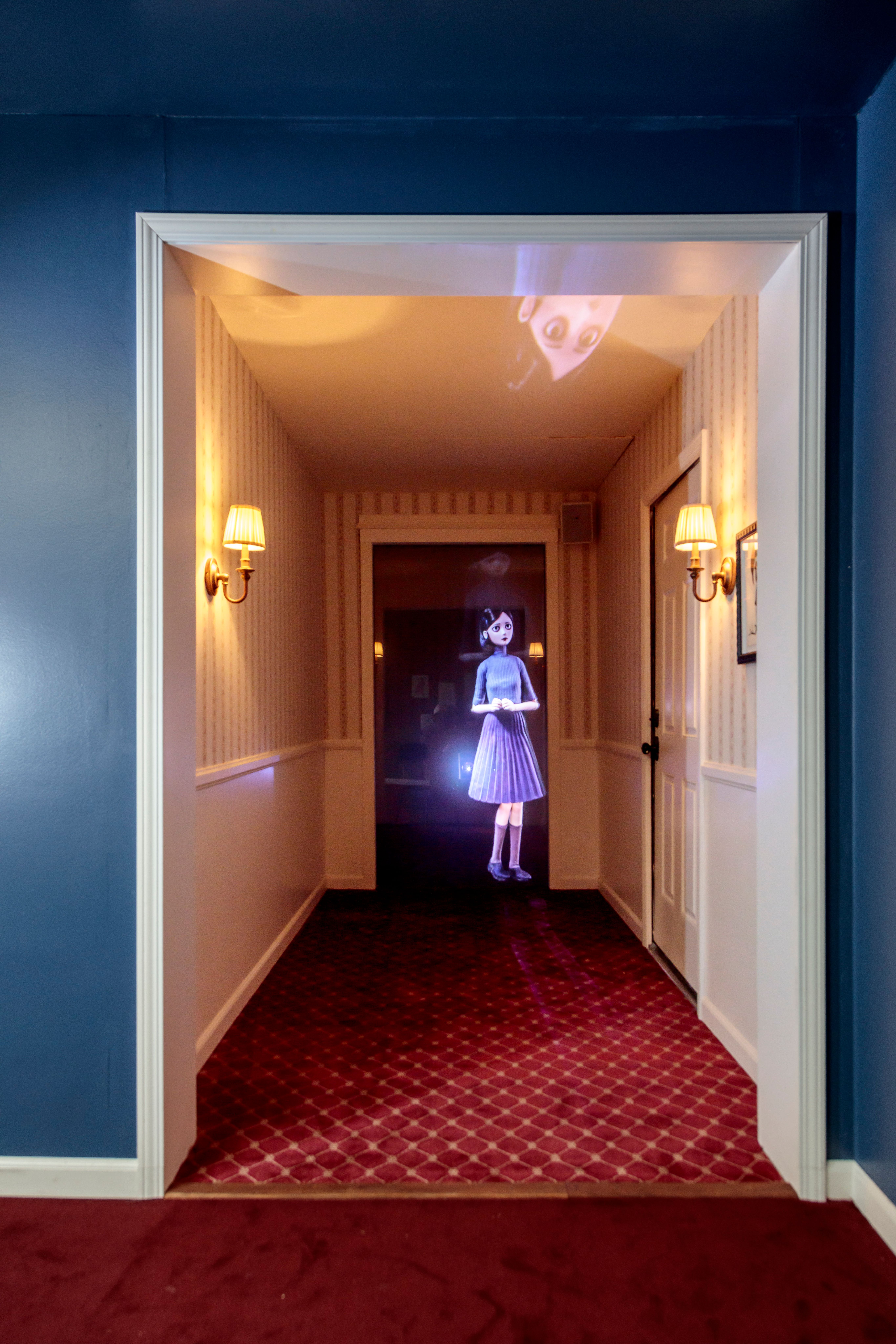 Projected animated girl stands in a warmly lit, striped hallway with red patterned carpet.
