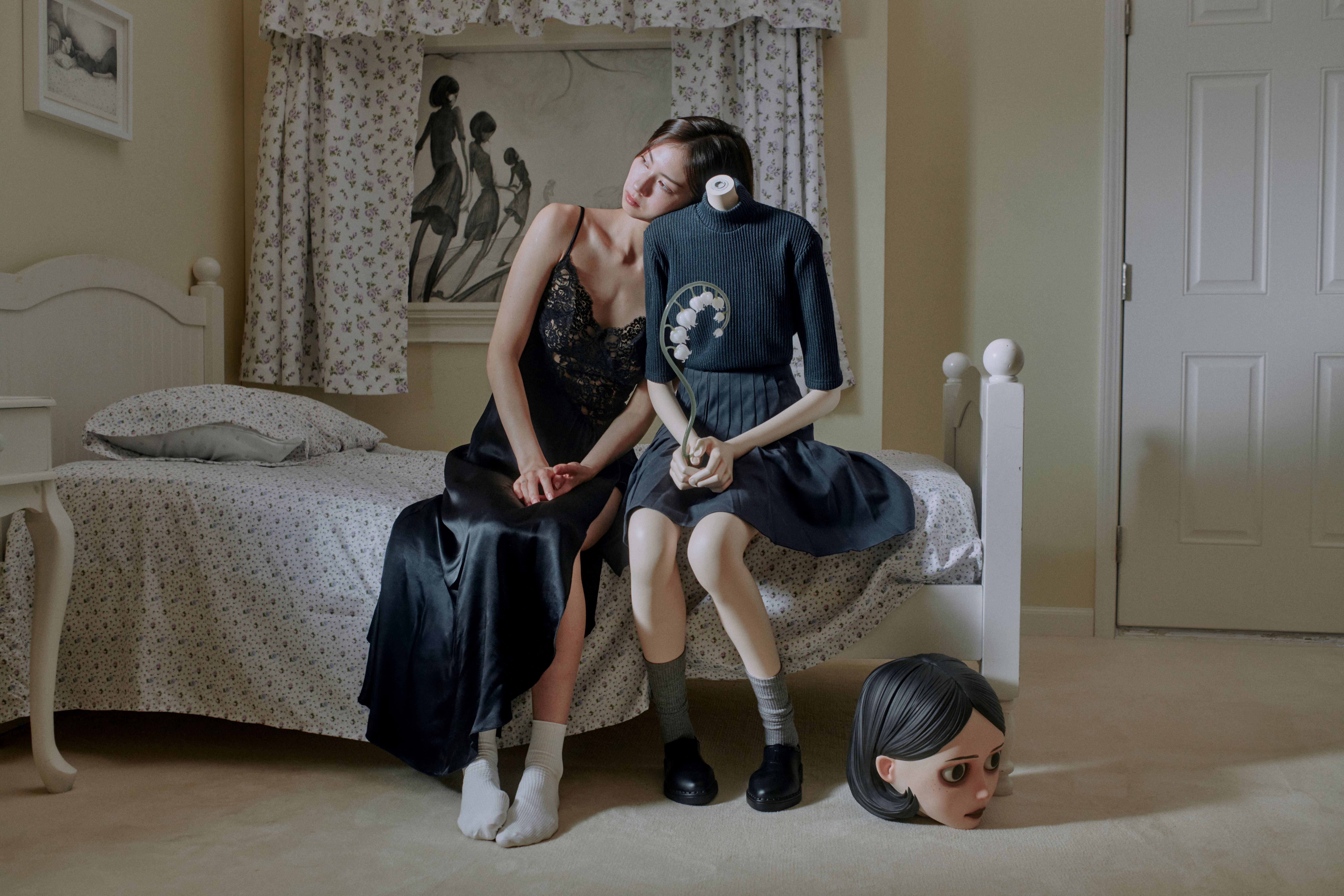 Woman in a black dress rests against a headless mannequin holding flowers, with a doll head on the floor.