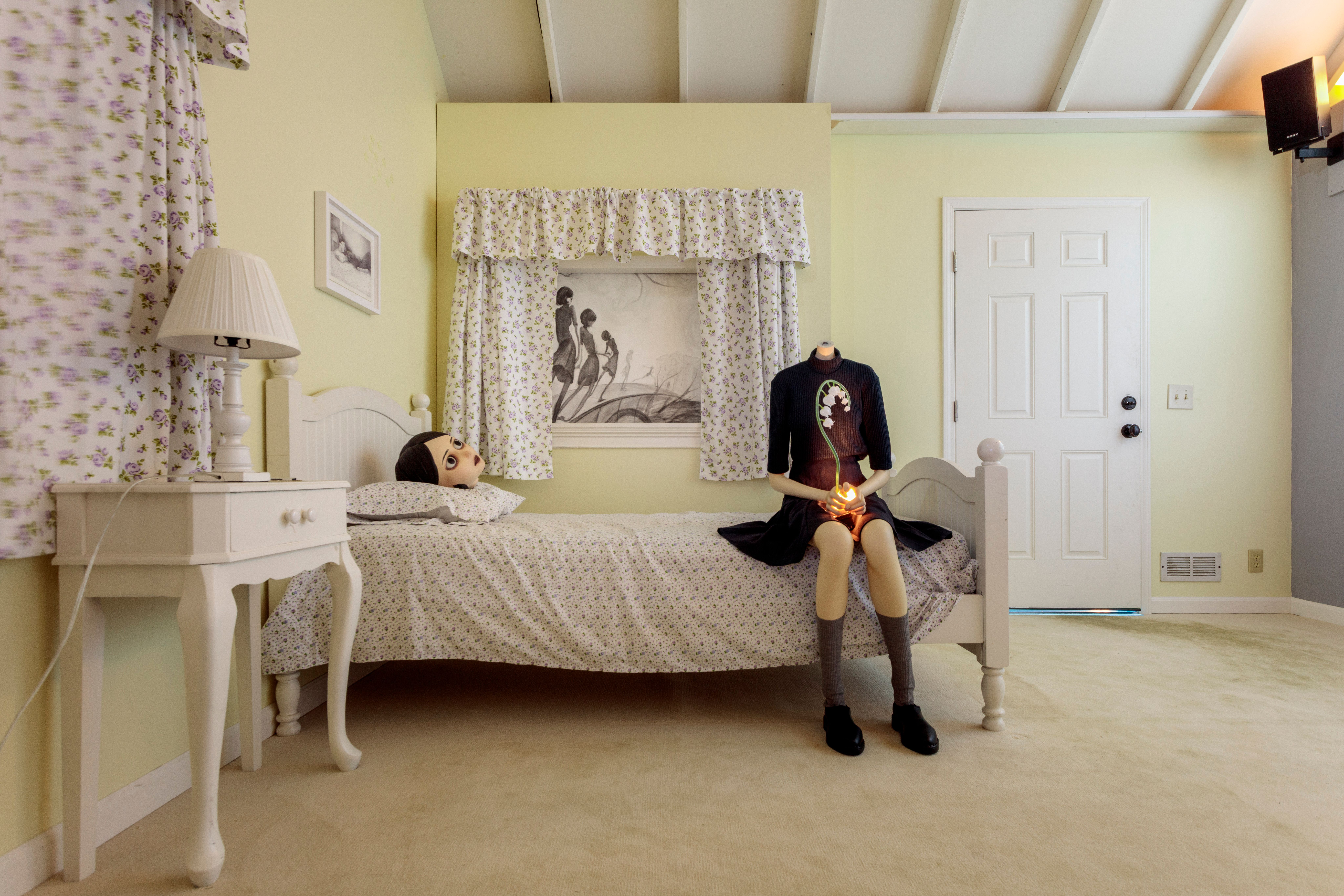 Surreal headless figure holding a glowing flower sits on a bed beside an oversized doll head in a floral bedroom.