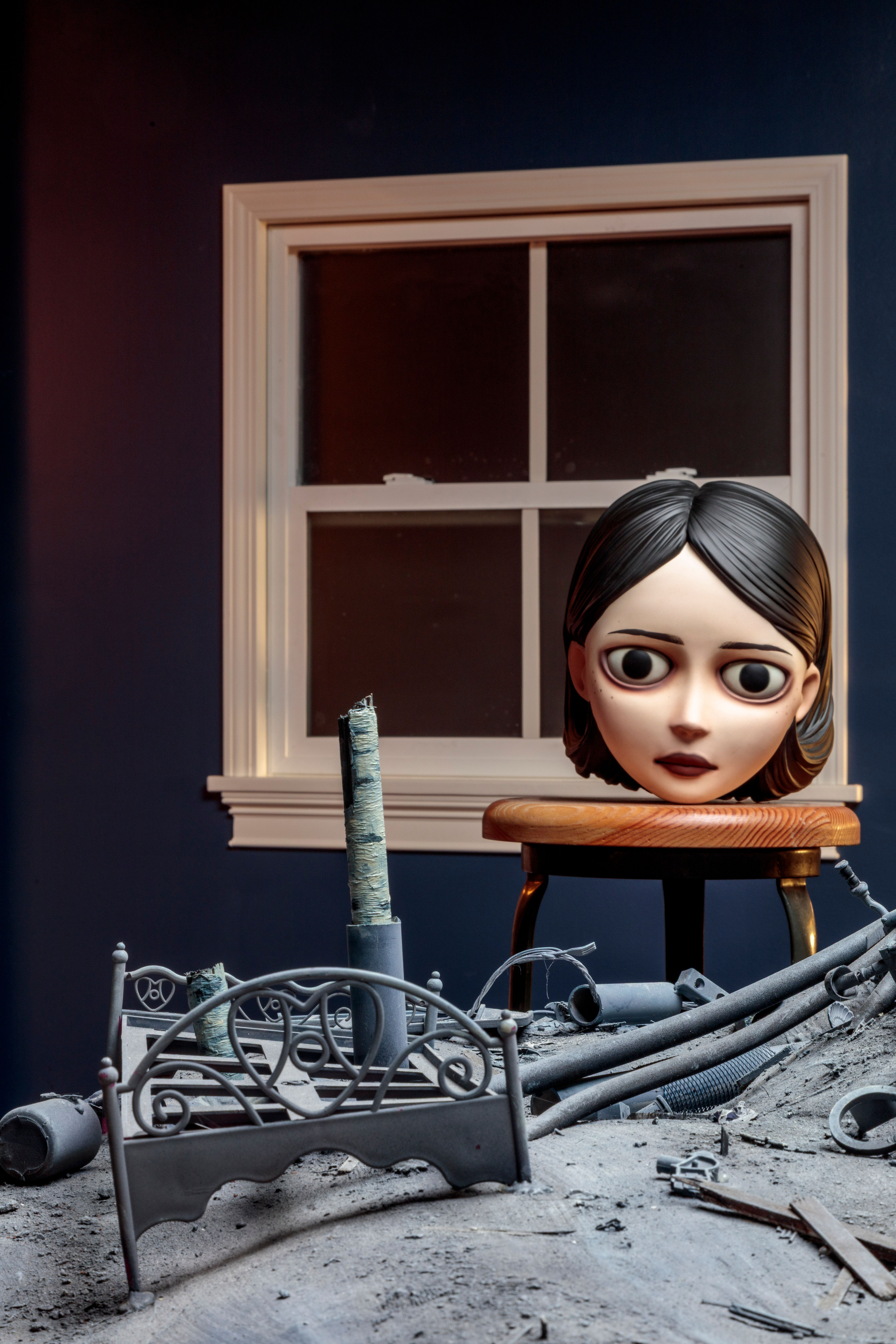 Surreal 3D scene of a large doll head on a stool amid a dusty miniature bed frame and scattered debris.