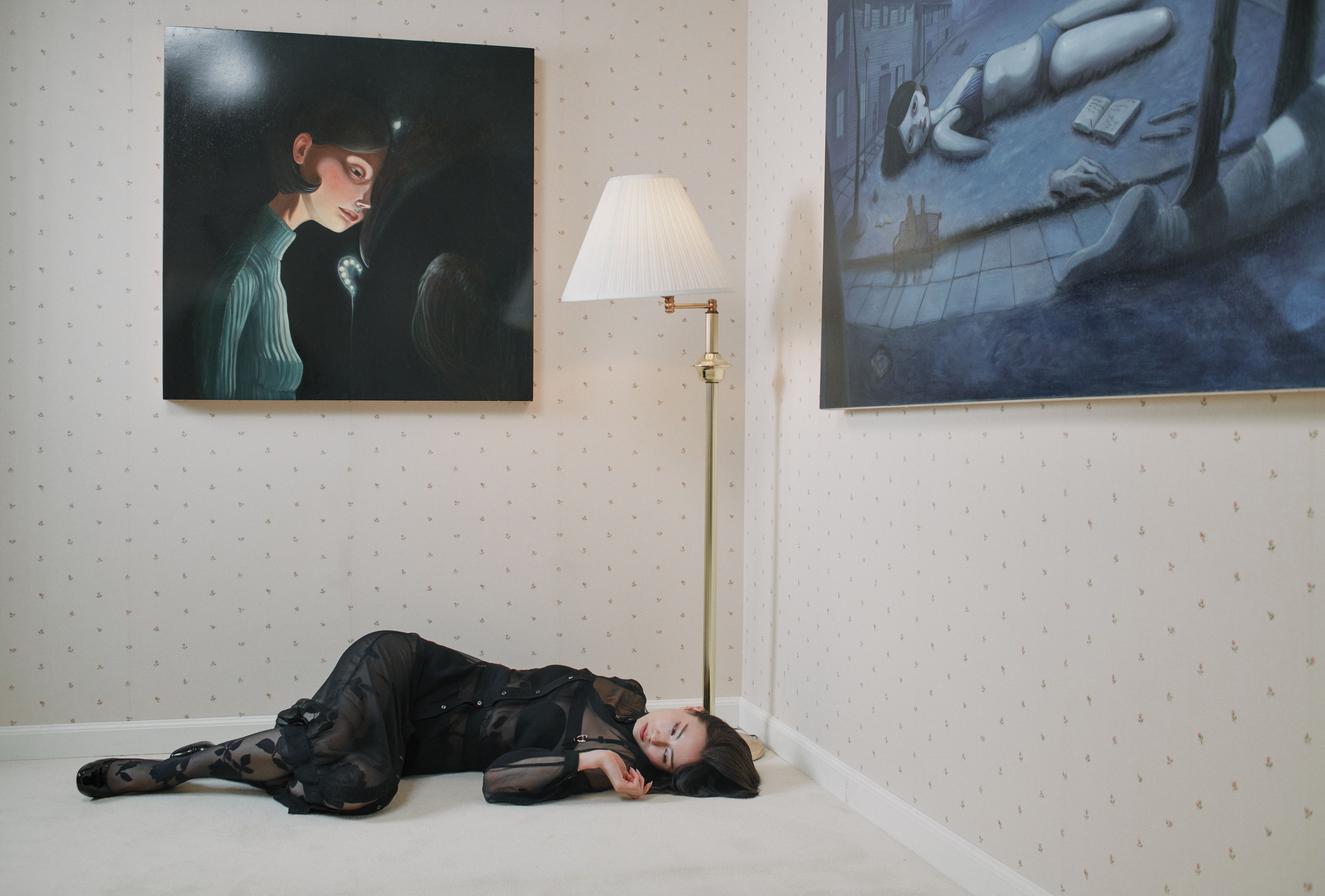 Woman in a sheer black dress lies beneath a floor lamp between two surreal paintings.