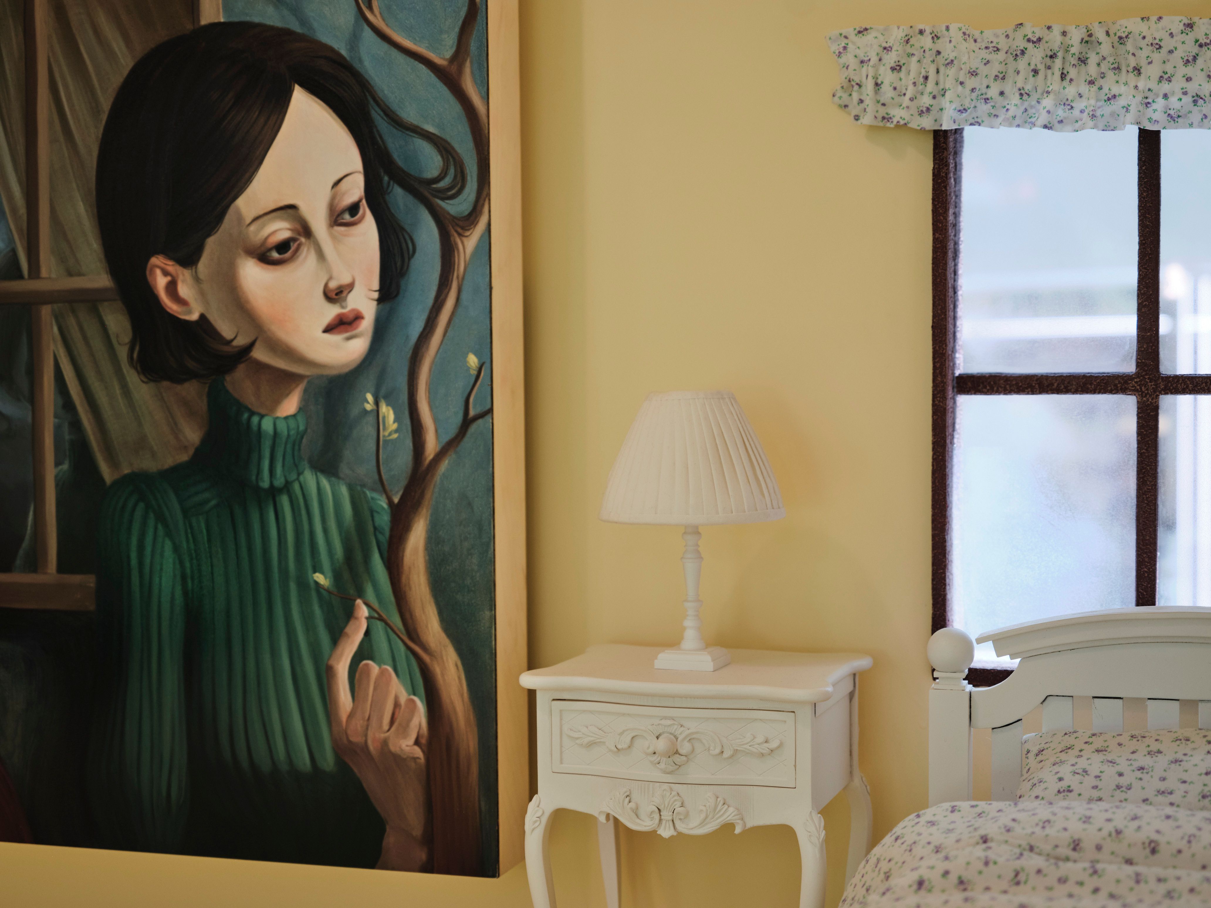 Surreal portrait painting beside a white nightstand and floral bed in a yellow bedroom.