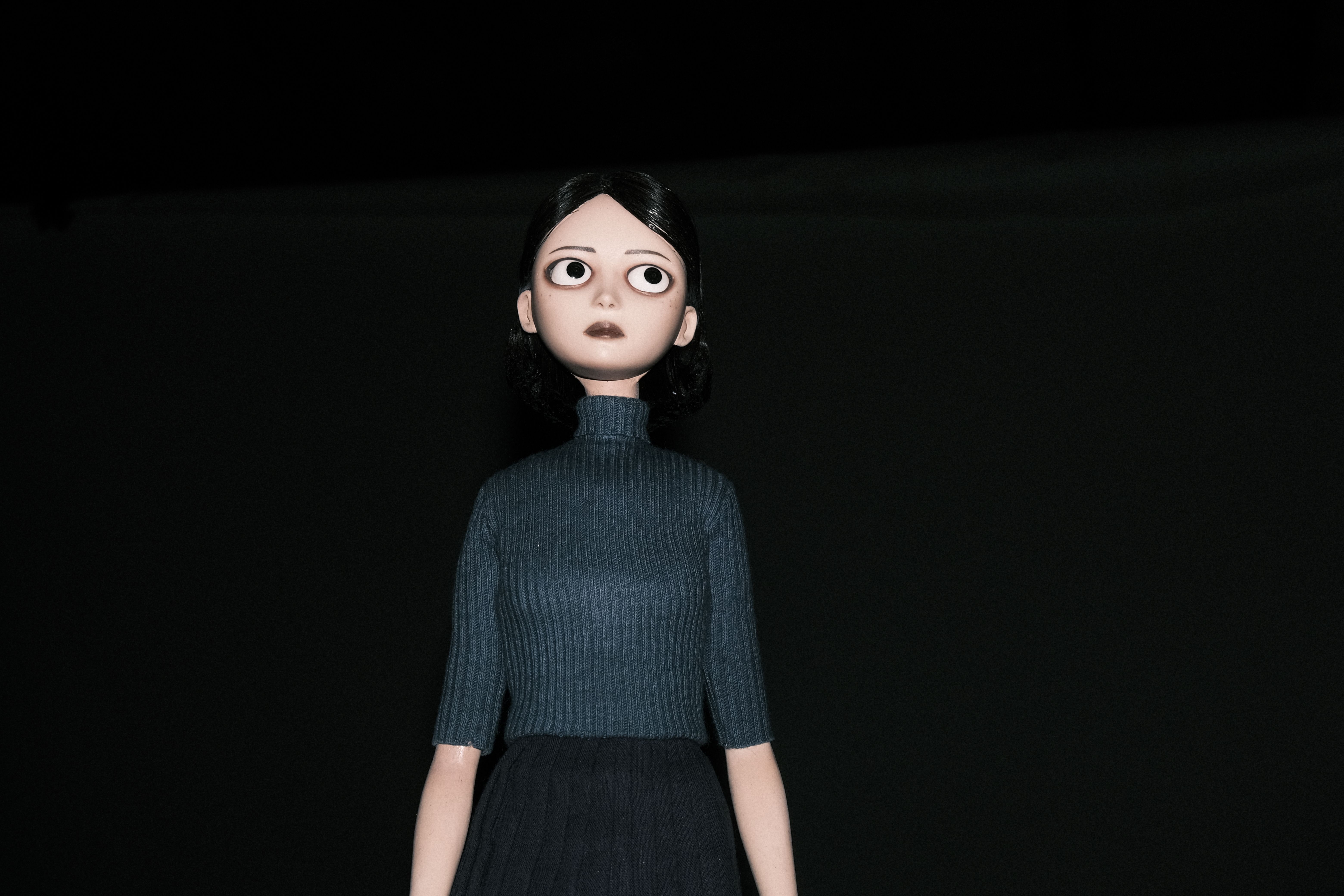 Stylized doll with large eyes, black bobbed hair, a gray turtleneck and black skirt against a dark background
