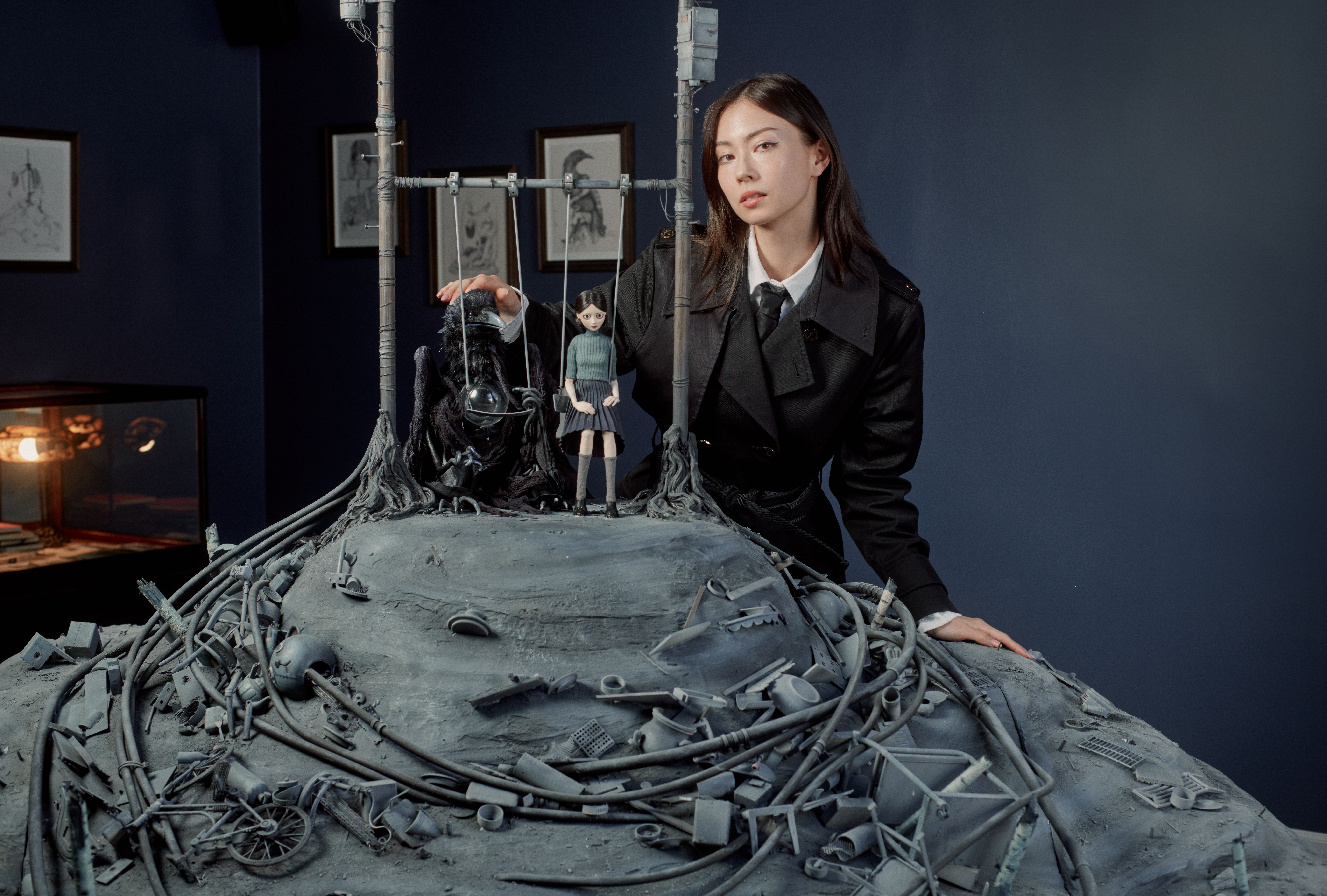 Woman in a black suit poses beside a gothic miniature set with a marionette, cables, and gray debris.