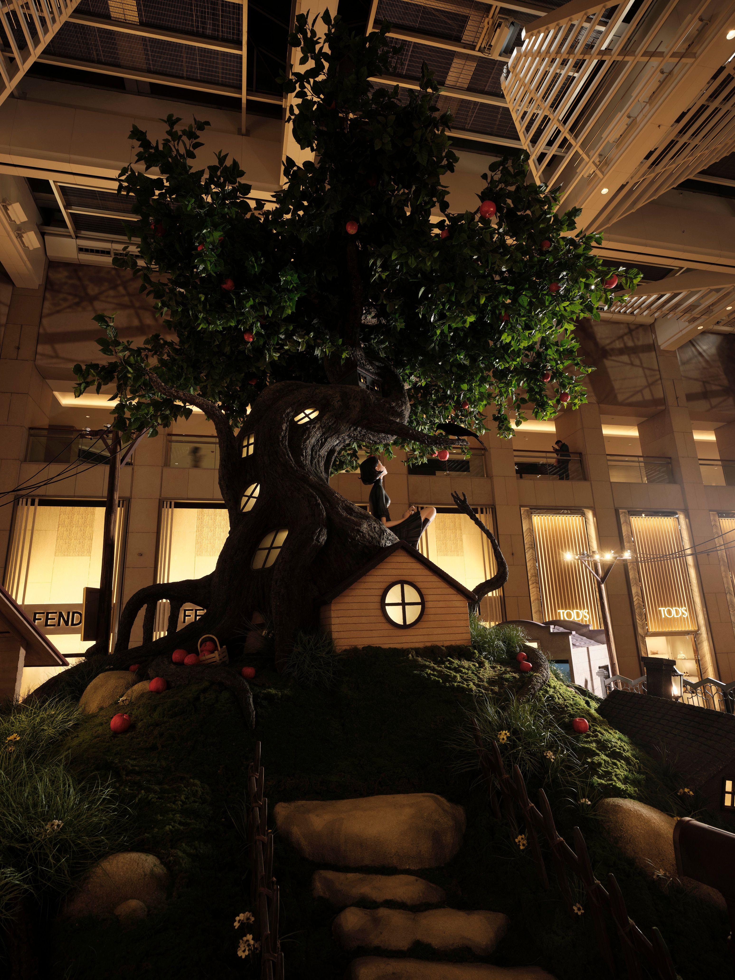 Whimsical indoor apple tree installation with glowing windows, a small house, and a seated figure