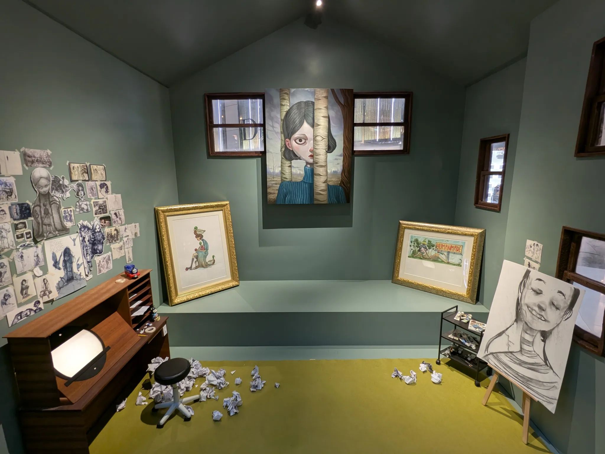 Artist's studio installation with framed paintings, sketches, easel, desk and crumpled paper on the floor.