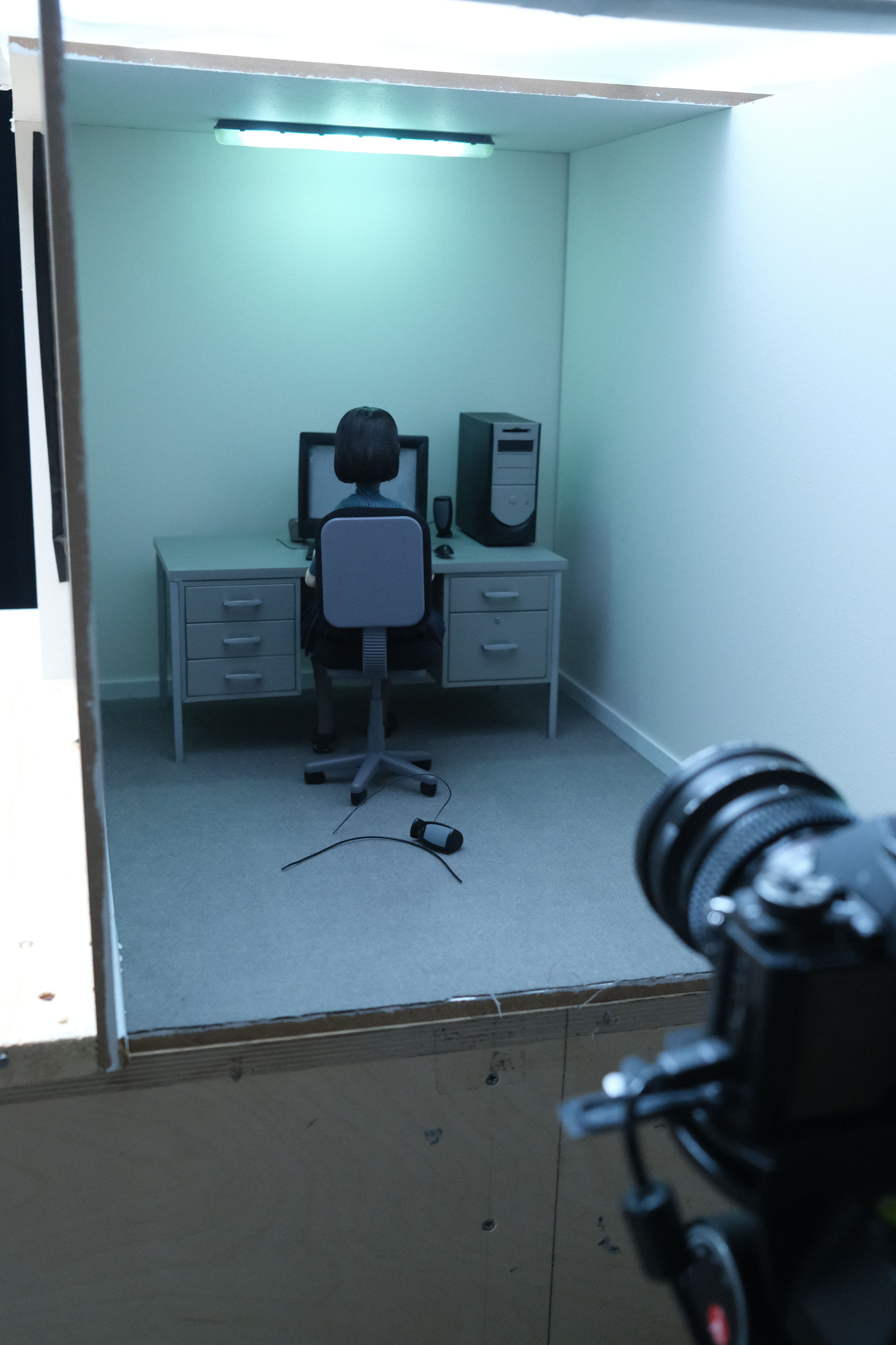 Miniature office set with a seated figure at a computer, photographed beside a visible camera.