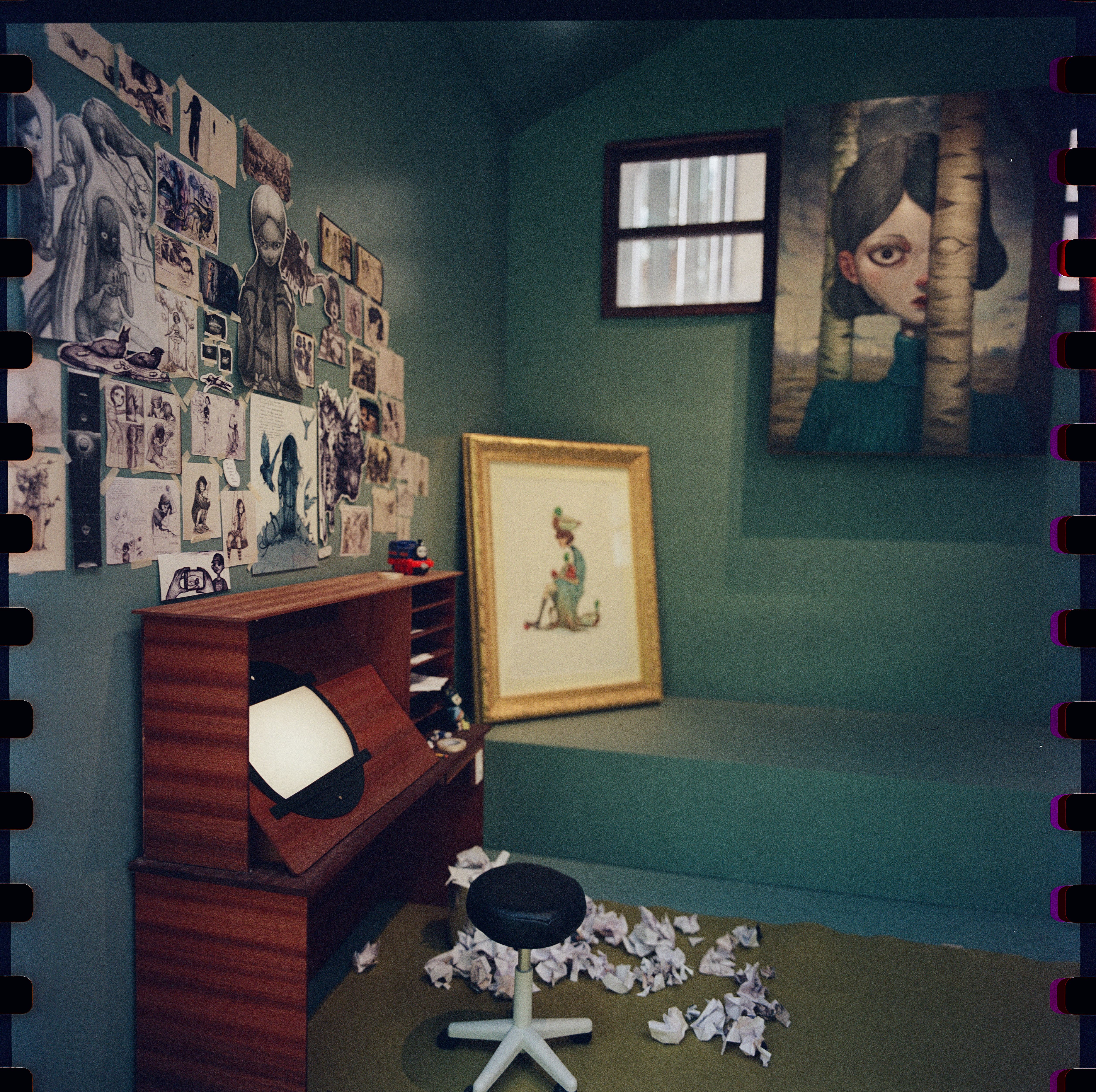 Teal artist's studio with sketch-covered wall, wooden desk, stool, paintings, and crumpled paper on the floor.