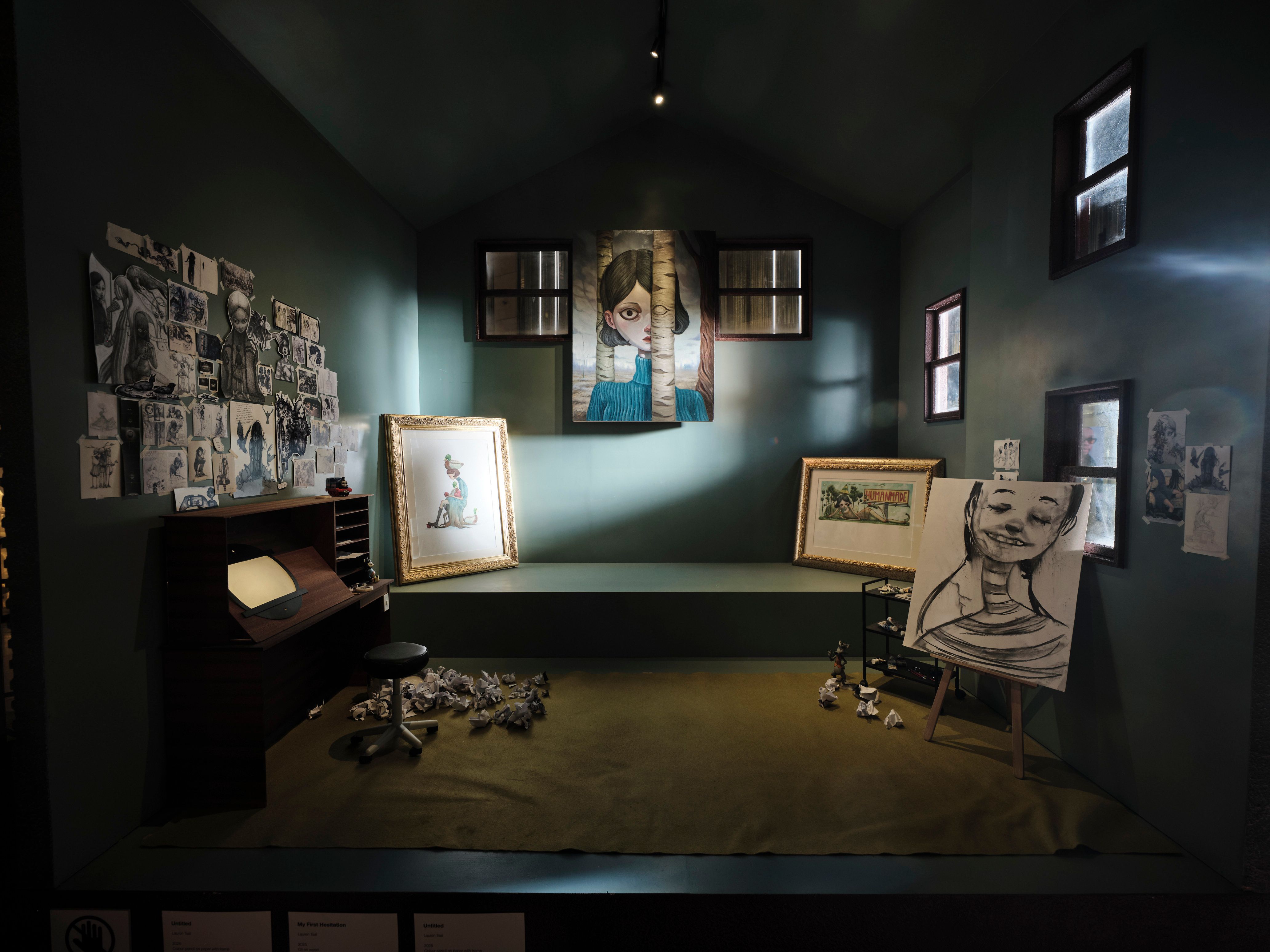Dimly lit artist's studio installation with eerie portraits, sketches, easel, desk and crumpled paper.