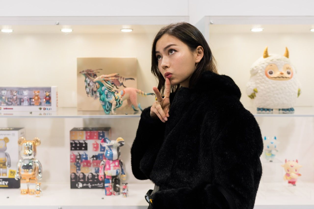Woman in a black fur coat poses with a peace sign before shelves of colorful designer toys.