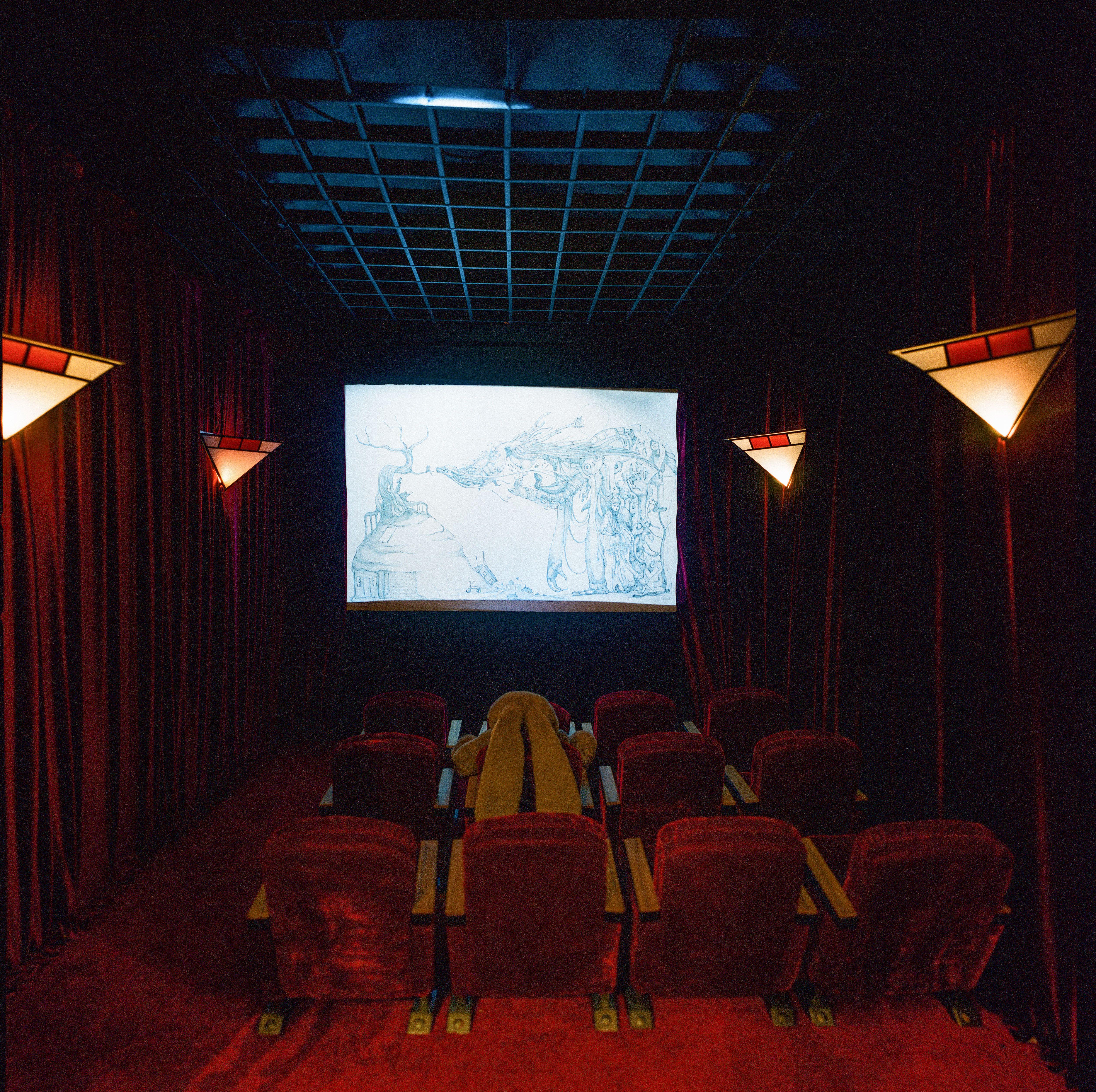 Small cinema with red velvet seats and curtains, wall sconces, and a sketch projected on the screen