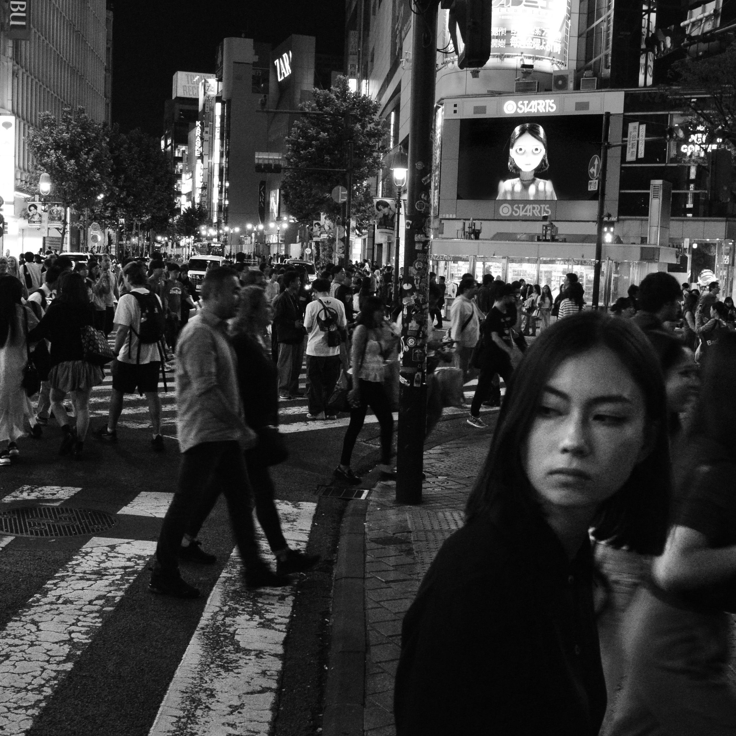 Black-and-white street photo of crowds crossing a neon-lit city intersection at night, with a woman in front.