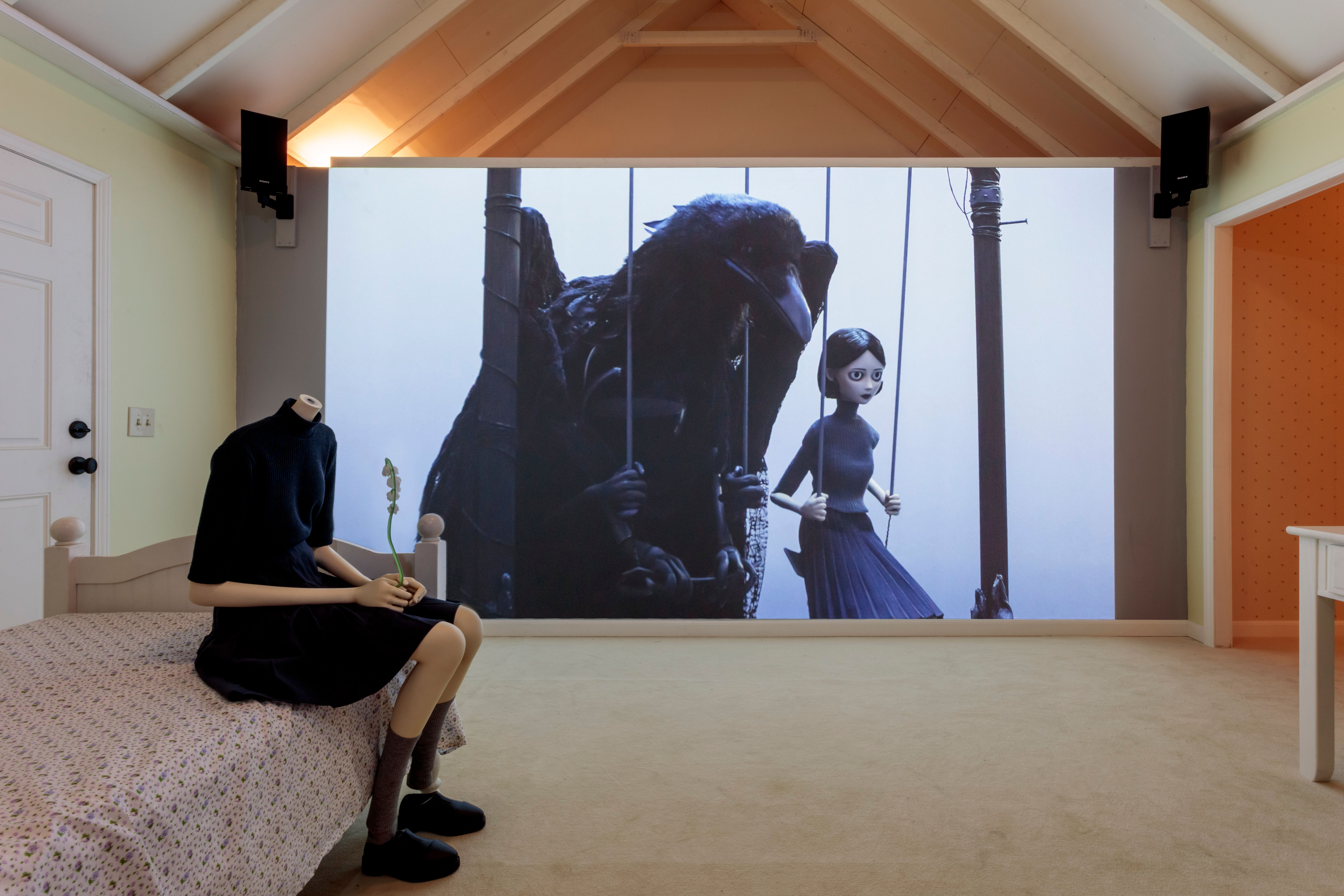 Headless mannequin holding a flower sits on a bed before a wall-sized projection of a gothic animated film.
