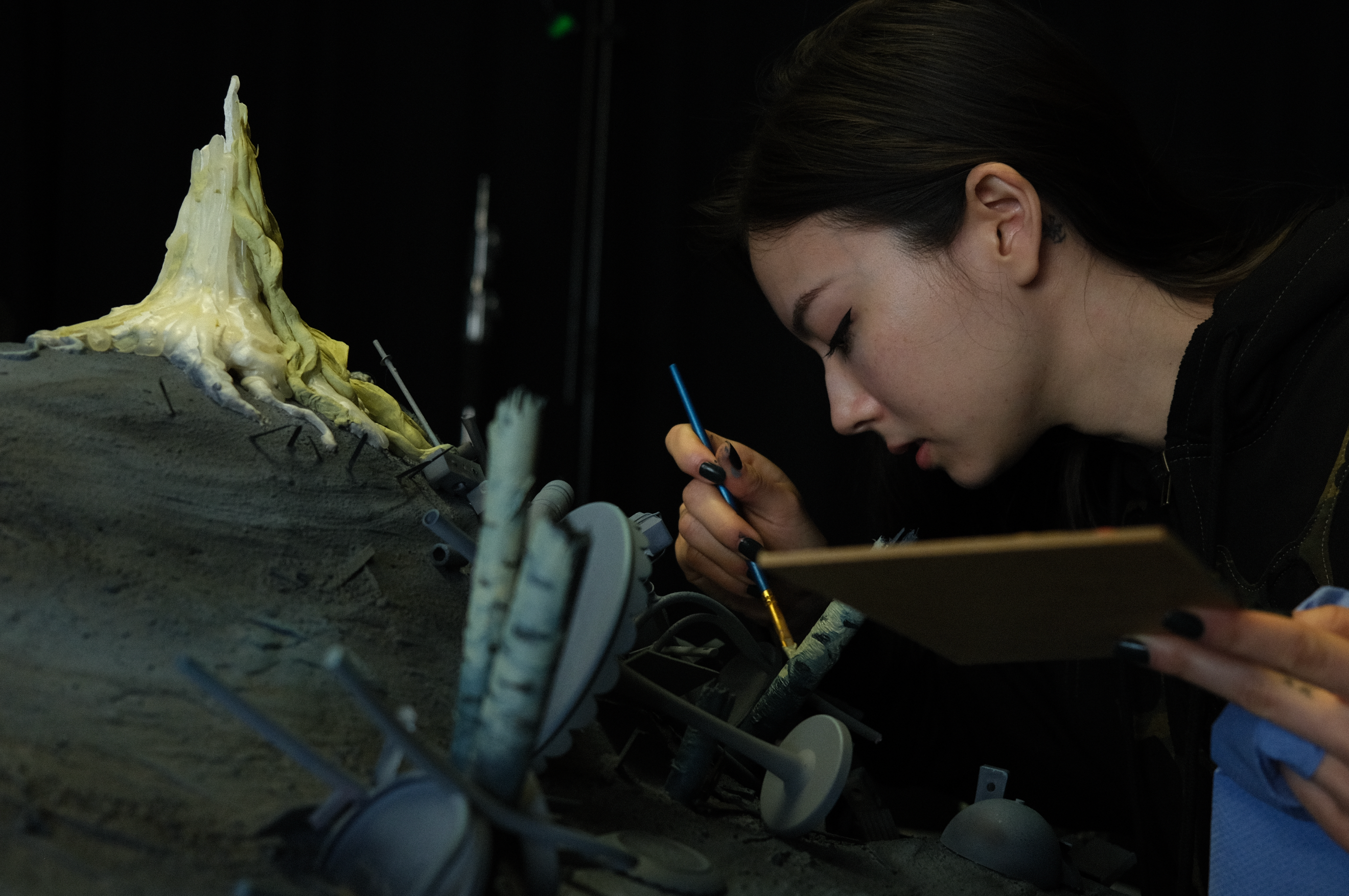 Artist uses a fine brush and palette to paint details on a large gray sculptural model.