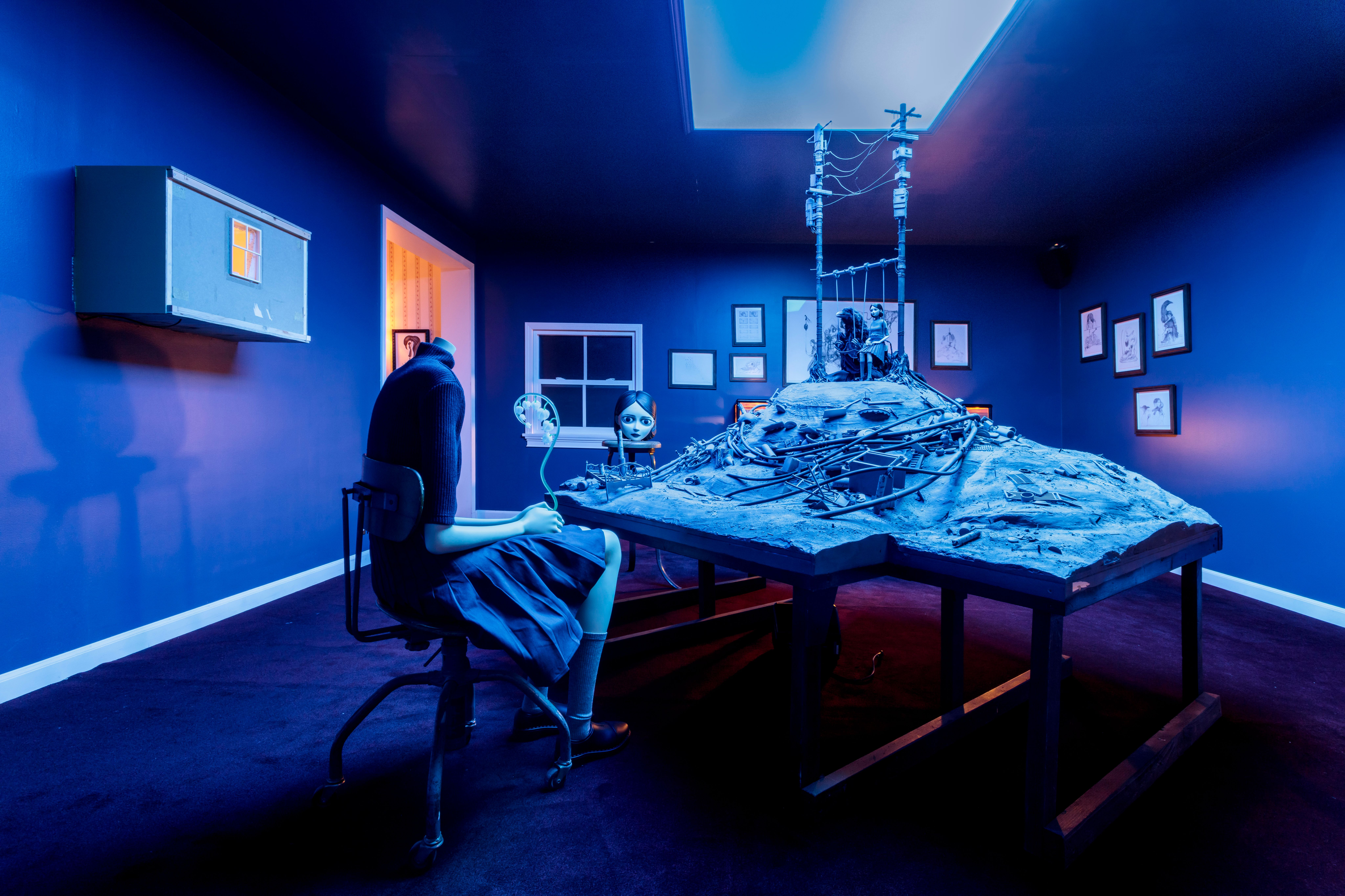 Surreal blue-lit installation with a seated mannequin facing a debris-covered table and miniature power lines.