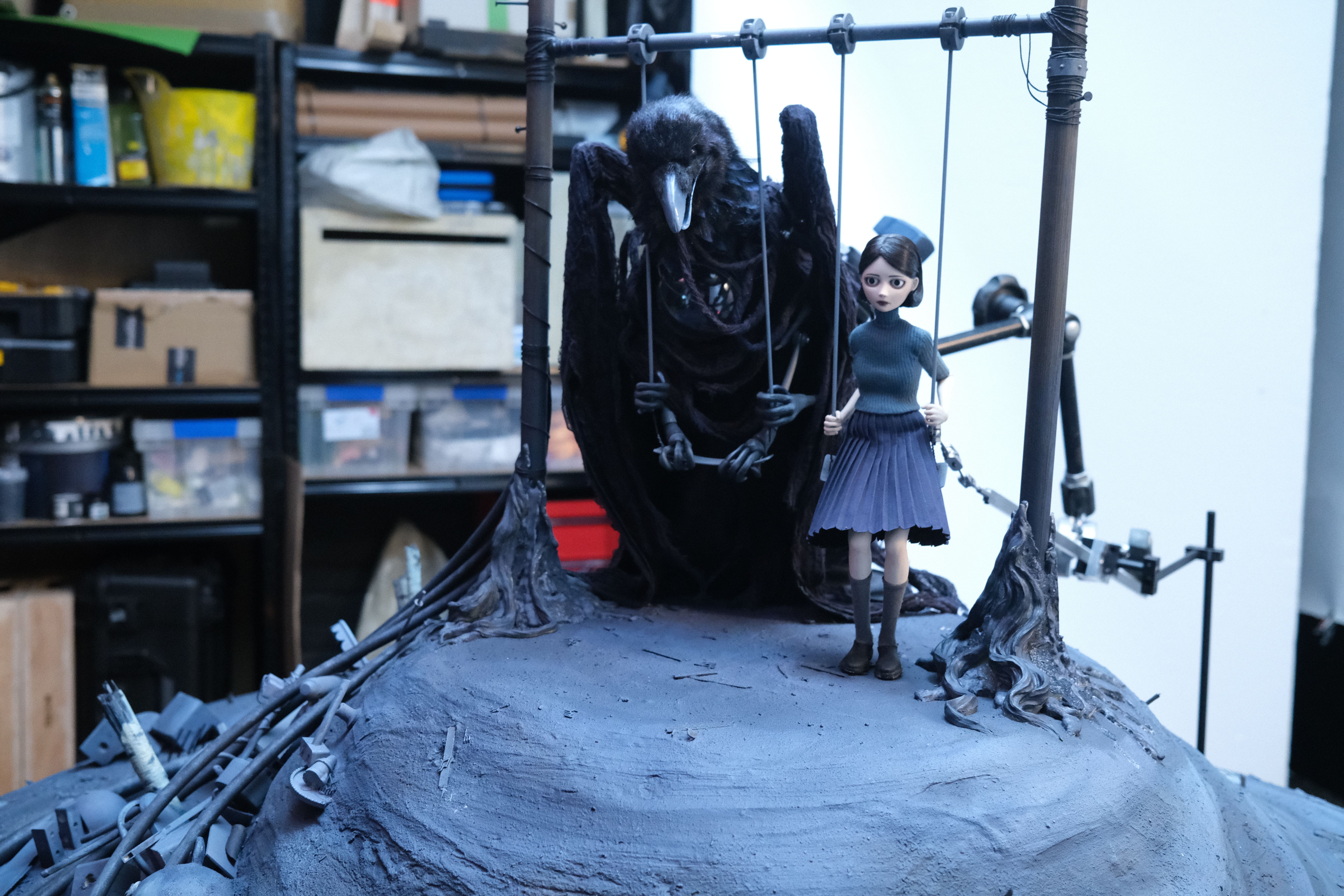 Gothic stop-motion set with a girl puppet beside a large black bird on a swing in a workshop.