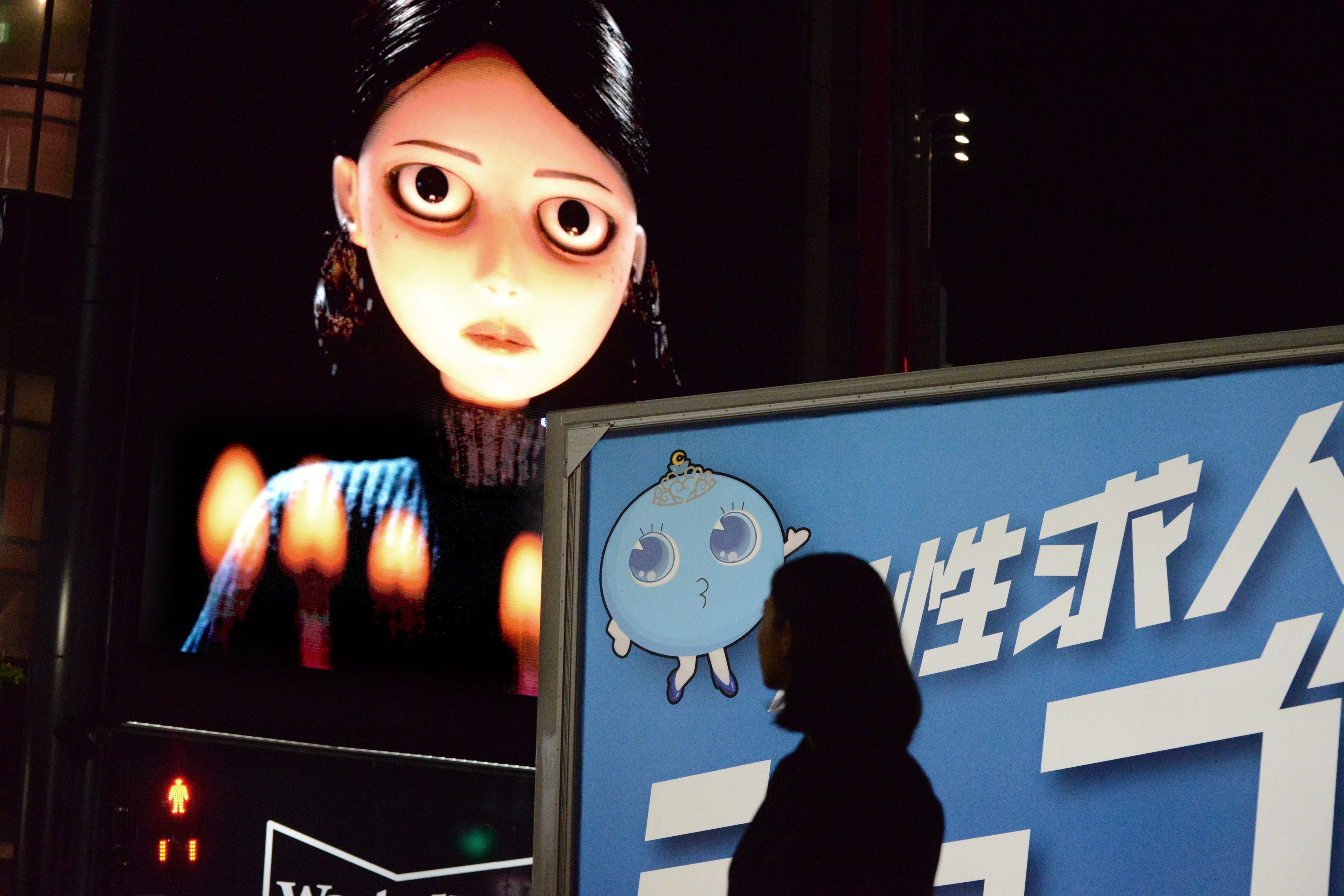 Silhouetted pedestrian passes a blue Japanese sign beneath a giant animated woman on a digital billboard.