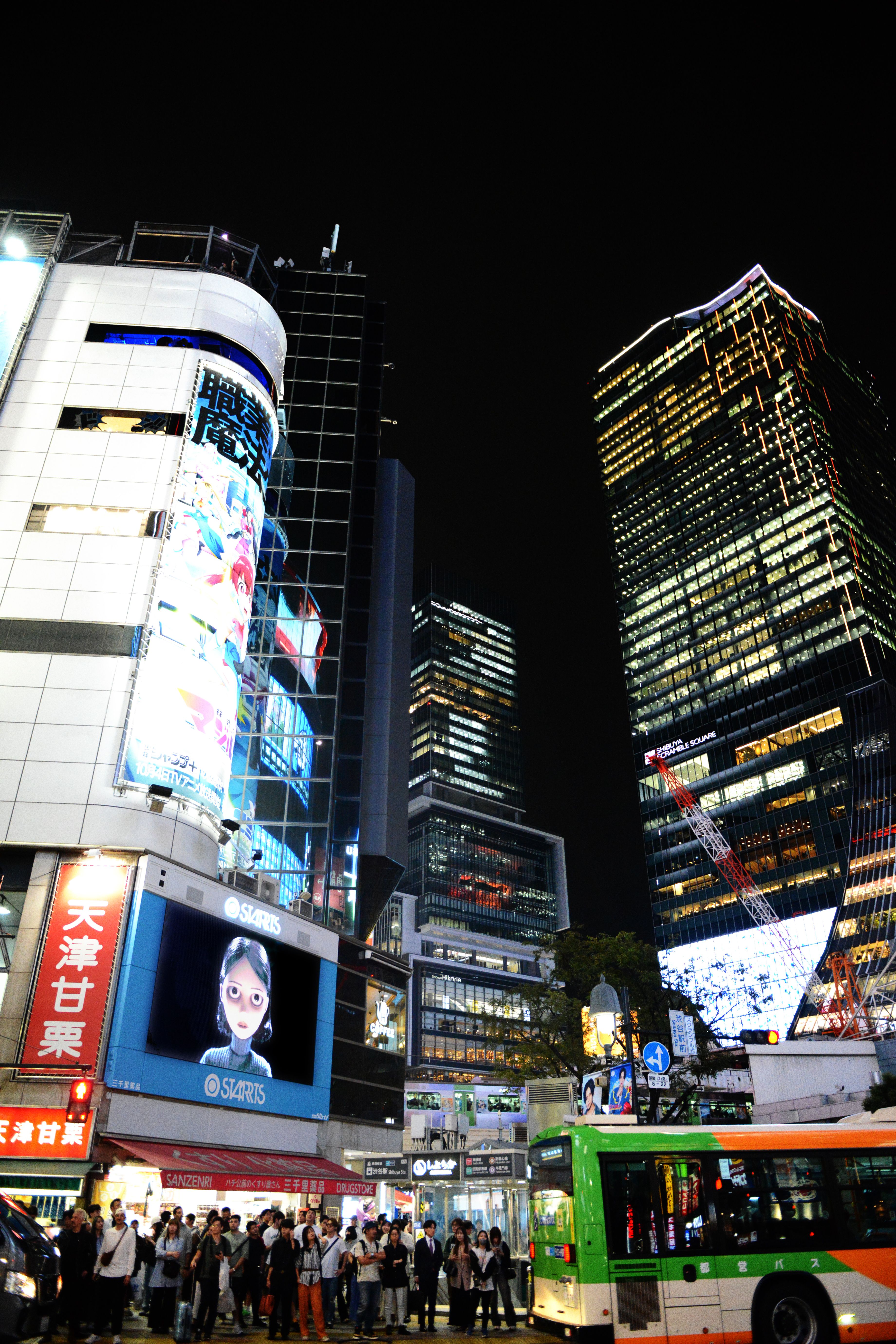 Crowds and a green bus beneath illuminated billboards and skyscrapers in Tokyo at night