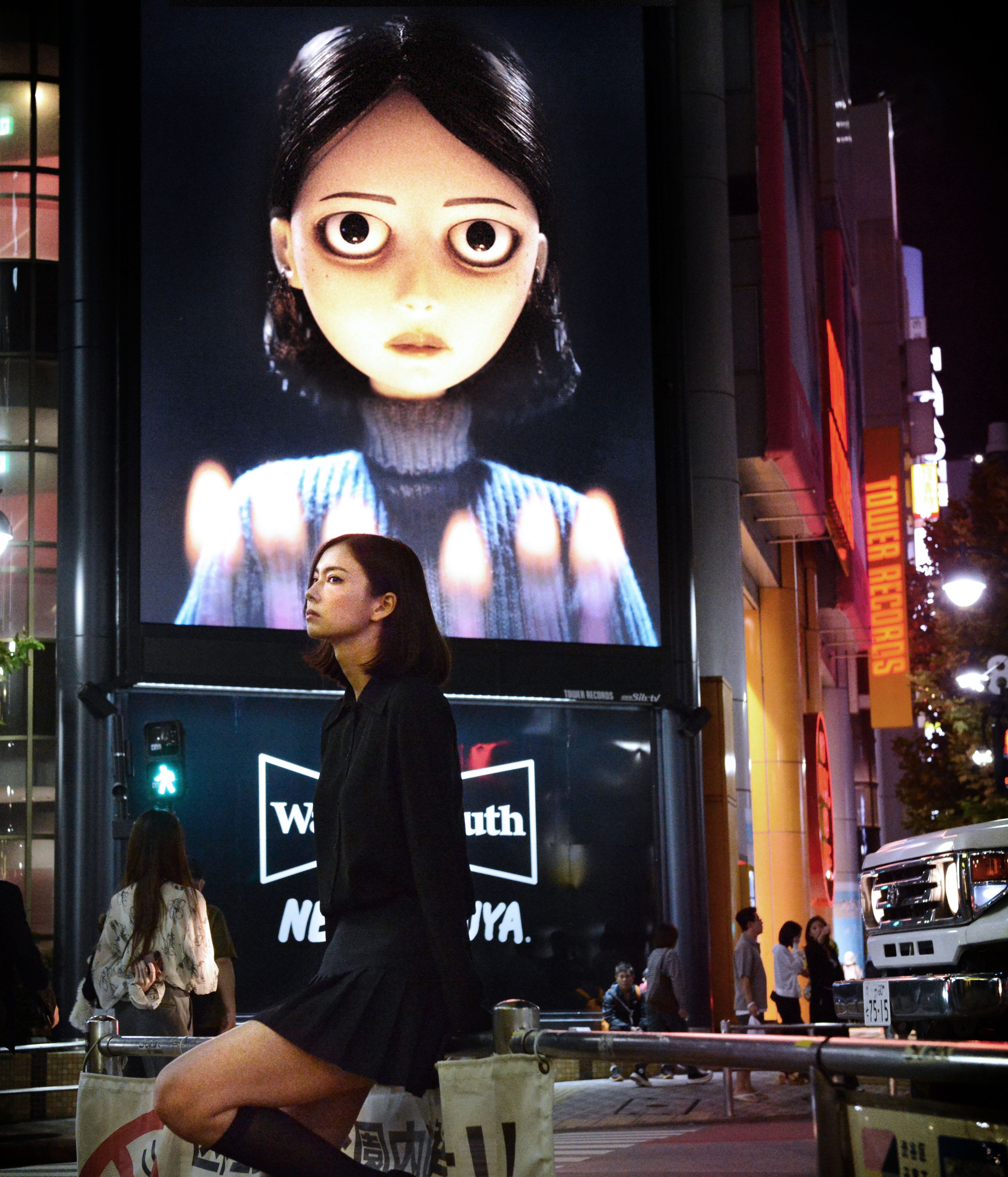 Woman in black poses on a neon-lit city street beneath a giant digital billboard of a wide-eyed animated girl.