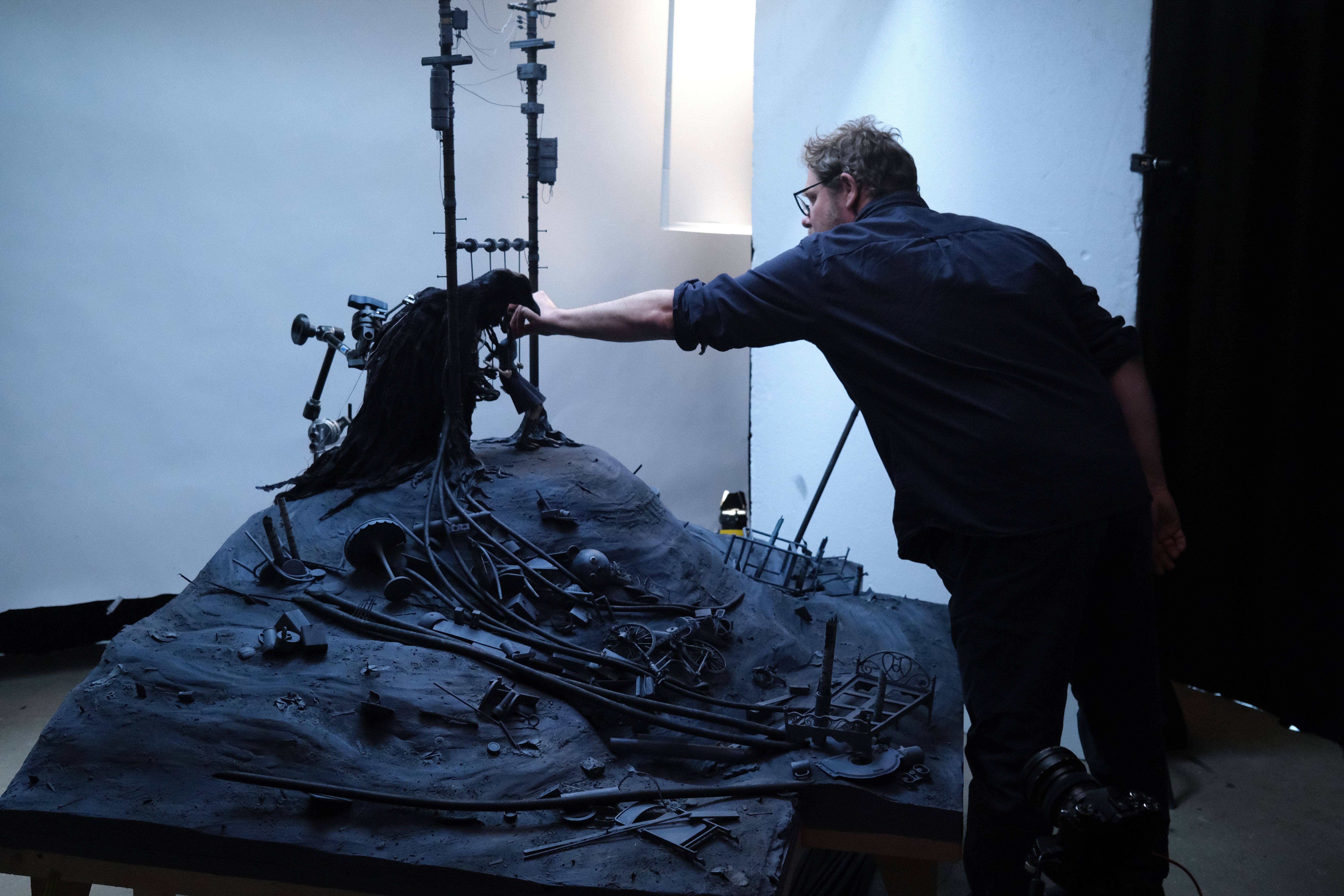 Artist adjusts a dark sculptural installation of tangled cables, poles and mechanical debris on a table.