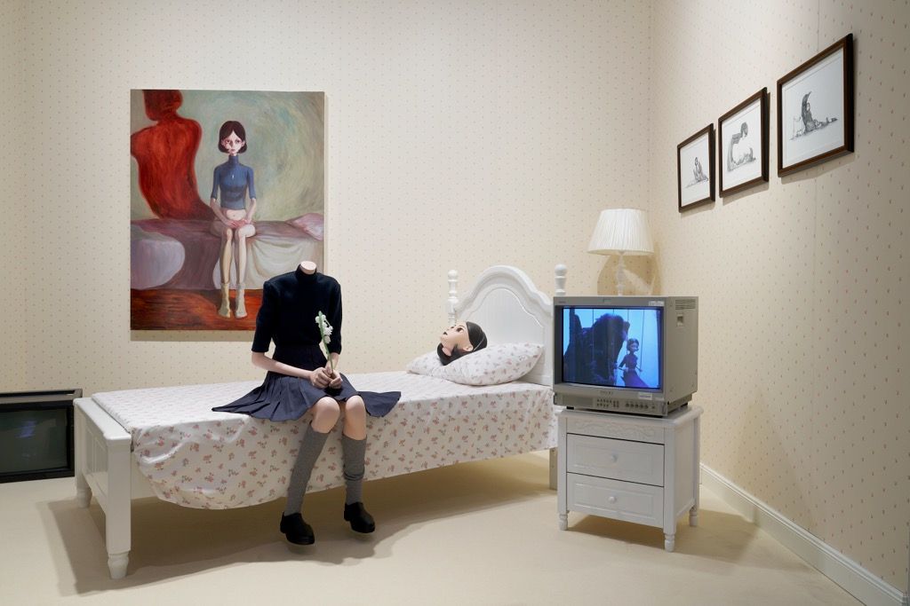 Surreal bedroom installation with a headless schoolgirl figure holding a flower beside a detached head and TV.