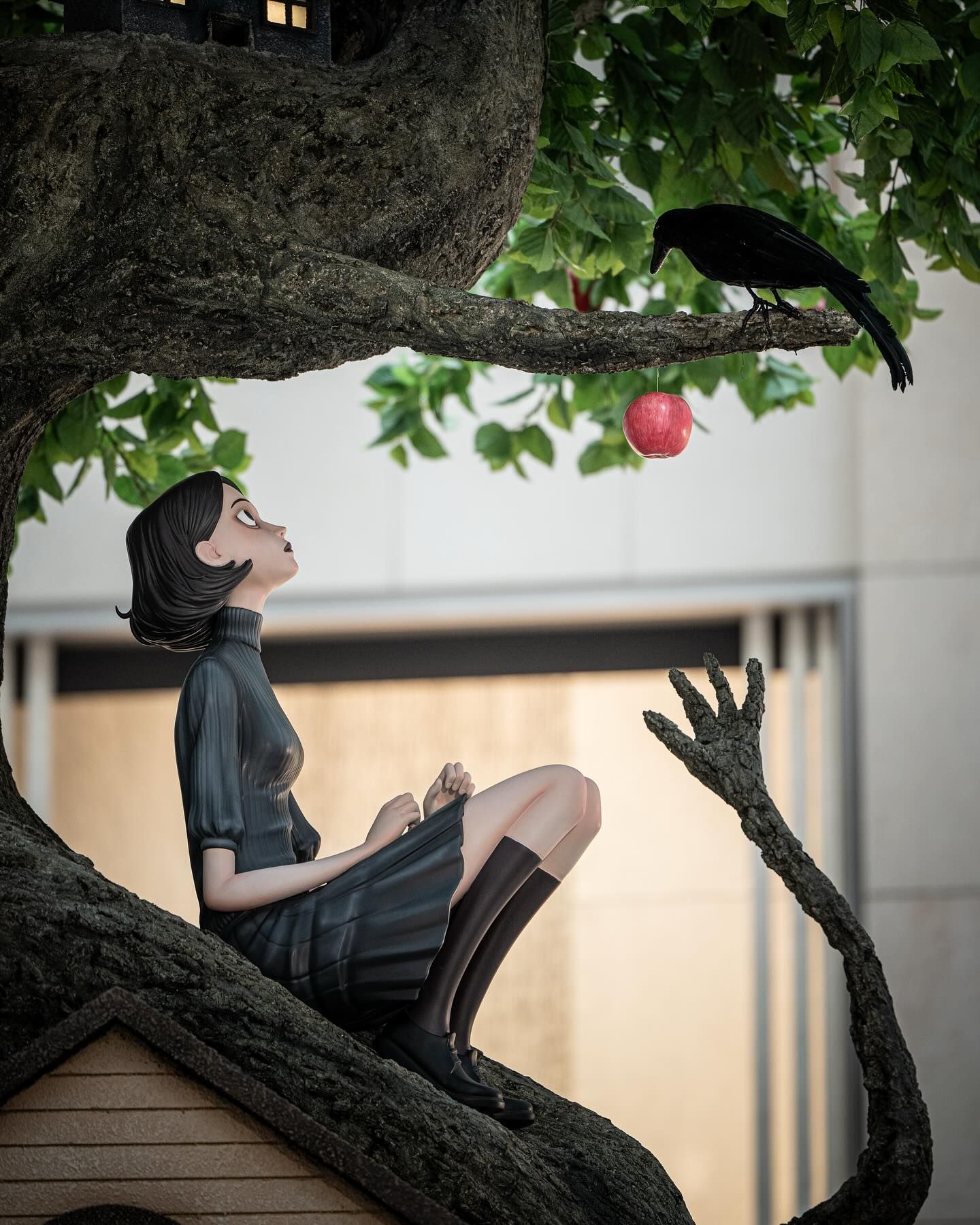 Sculpture of a gothic girl seated in a tree, looking up at a black crow and a hanging red apple