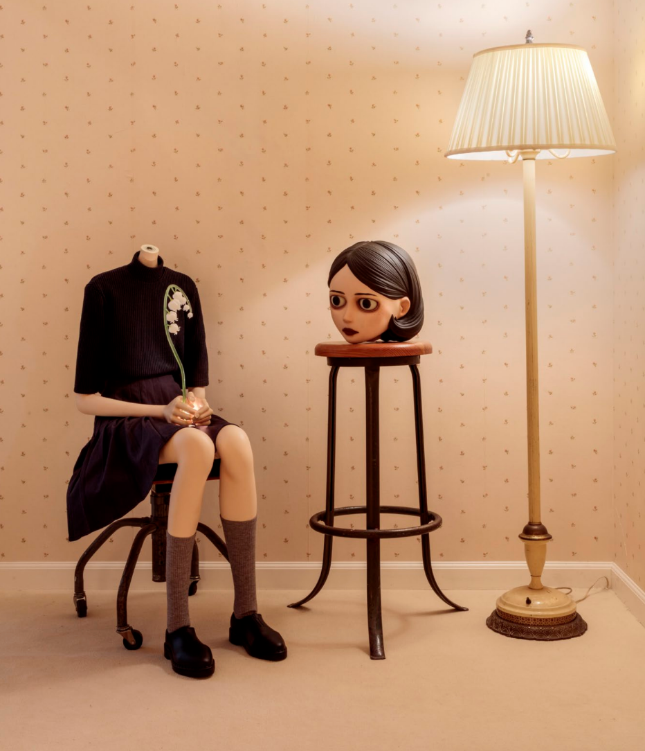 Surreal sculpture of a headless seated girl holding a flower, her head on a stool beside a floor lamp