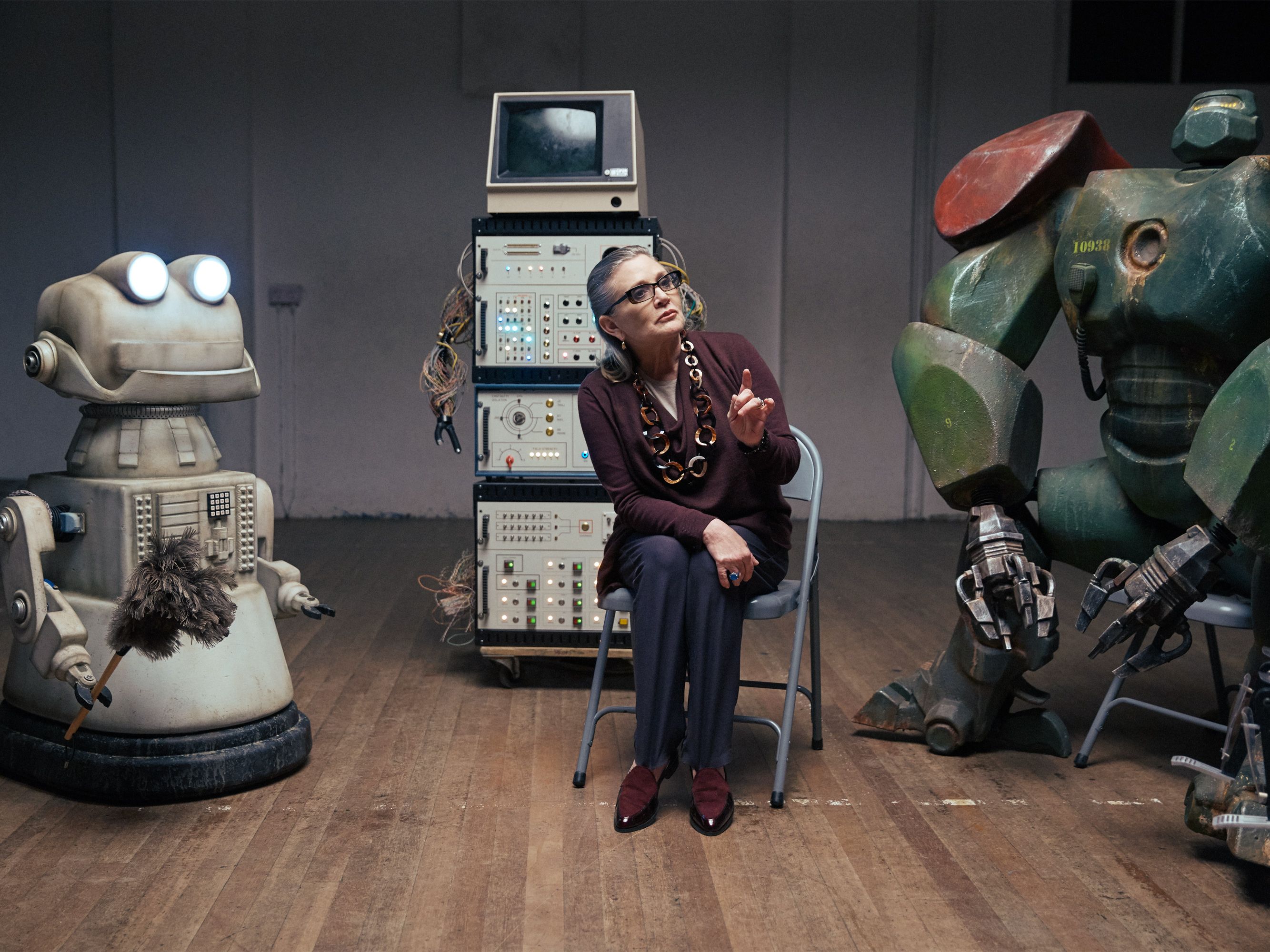 A woman sits in a room with two robots, one with a duster, and a large control panel in the background.
