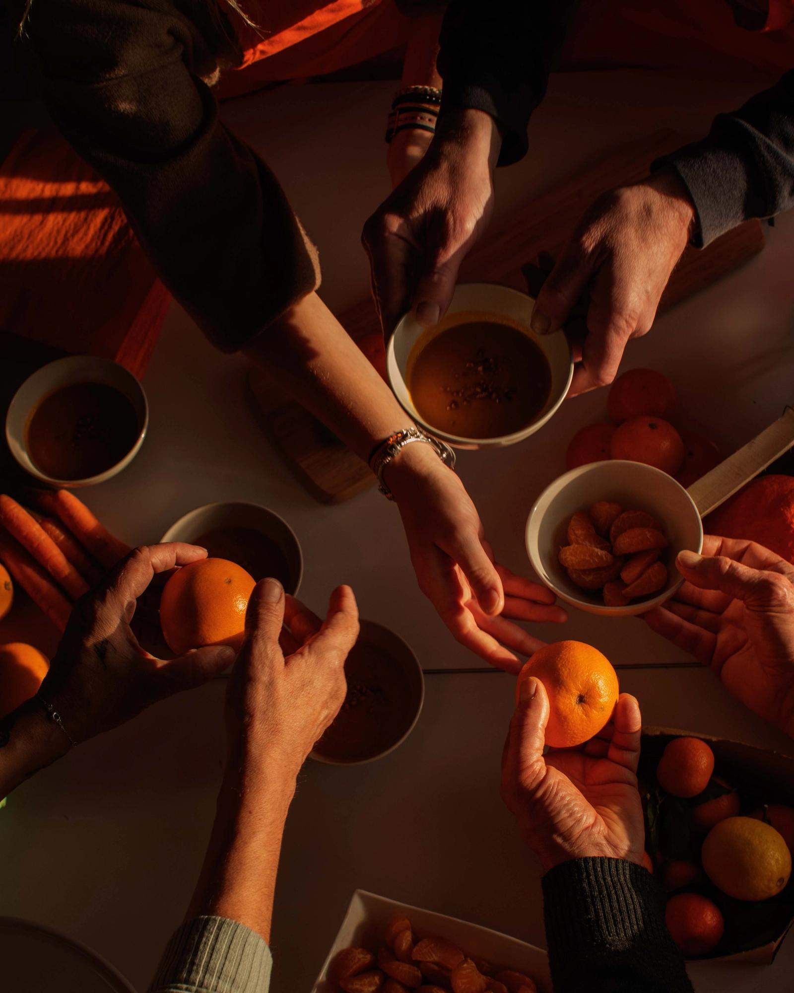 A group of people are gathered around a table, holding cups of coffee and oranges, with a few oranges and cups scattered around the table.