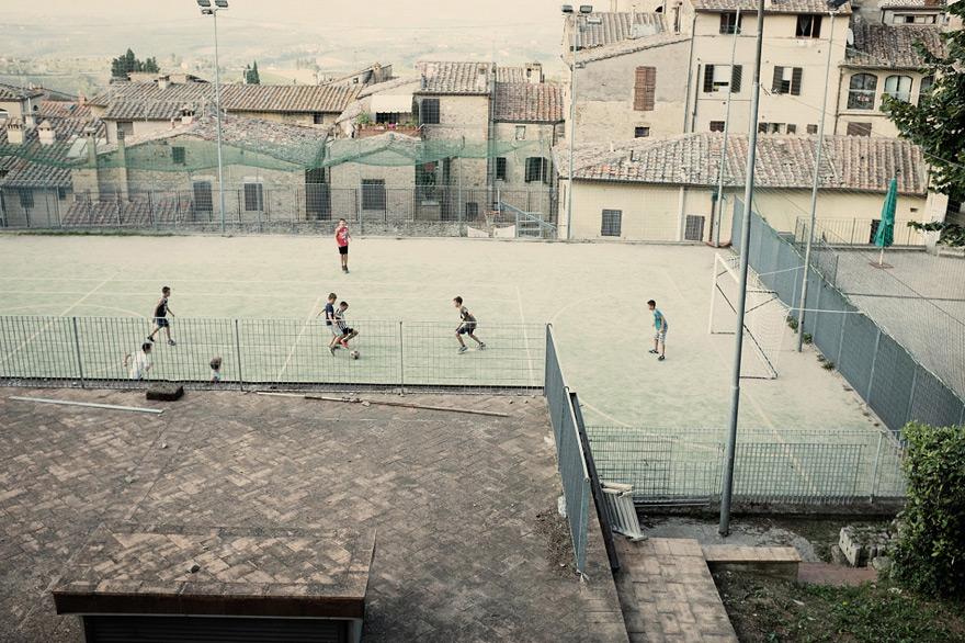 A group of children playing soccer in a small town's outdoor court, with a few buildings and a hazy sky in the background.