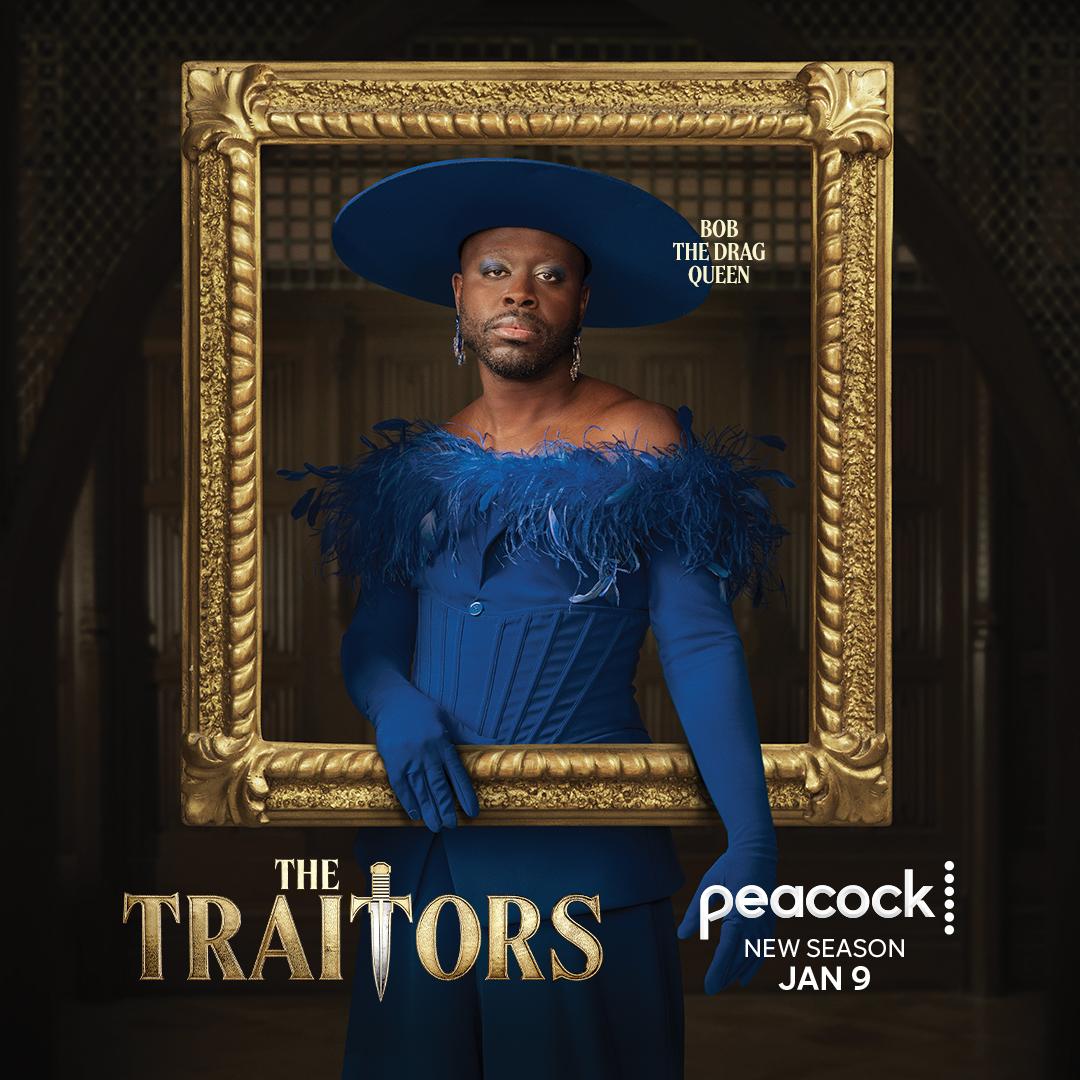 A person, likely a drag performer, dressed in a blue, feathered, corset-style top and a large, wide-brimmed blue hat, standing in front of a gold, ornate frame.