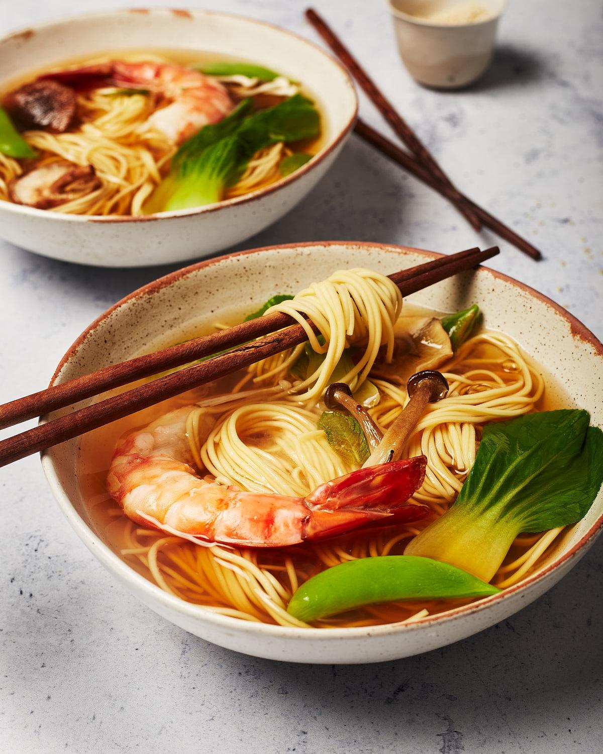 A bowl of noodles with a large shrimp, vegetables, and noodles in a broth, served with chopsticks.