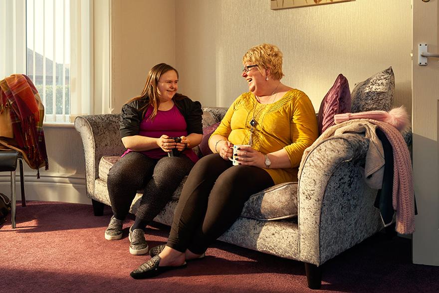 Two women sitting on a couch, both holding cups and smiling at each other.