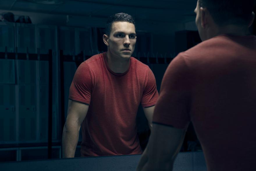 A man in a red t-shirt is looking at his reflection in a gym locker room mirror.