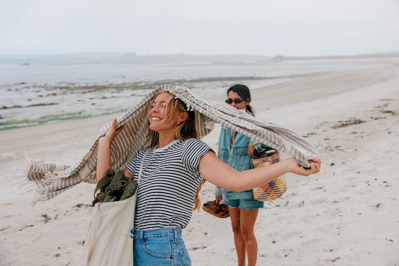 Two women on a beach, one holding a striped scarf over her head, the other in the background wearing a denim jacket and shorts, both carrying canvas bags.