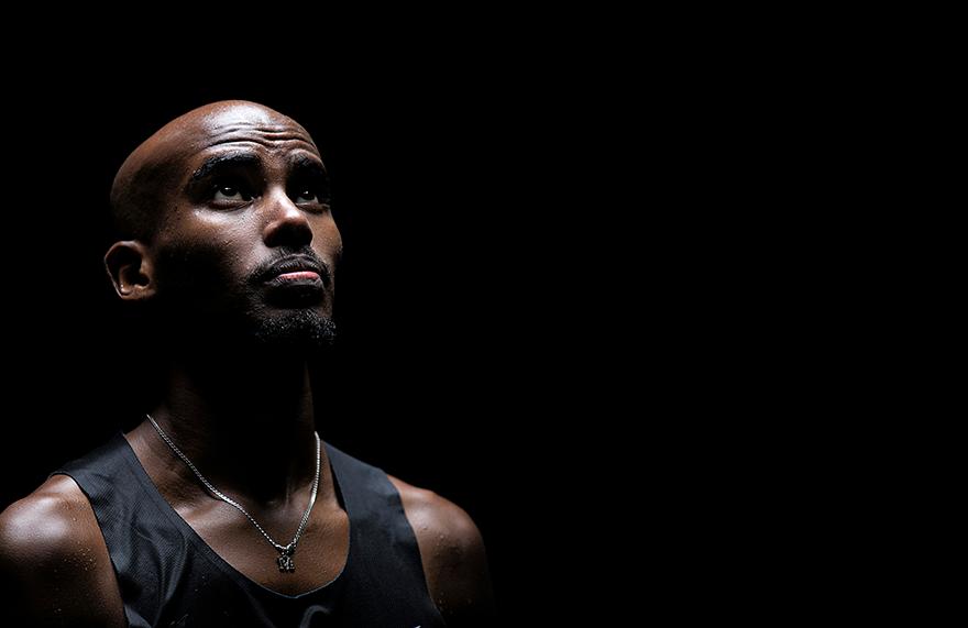 A close-up portrait of a bald, dark-skinned man with a strong, athletic build, wearing a black tank top and a silver chain with a small, cross-shaped pendant.
